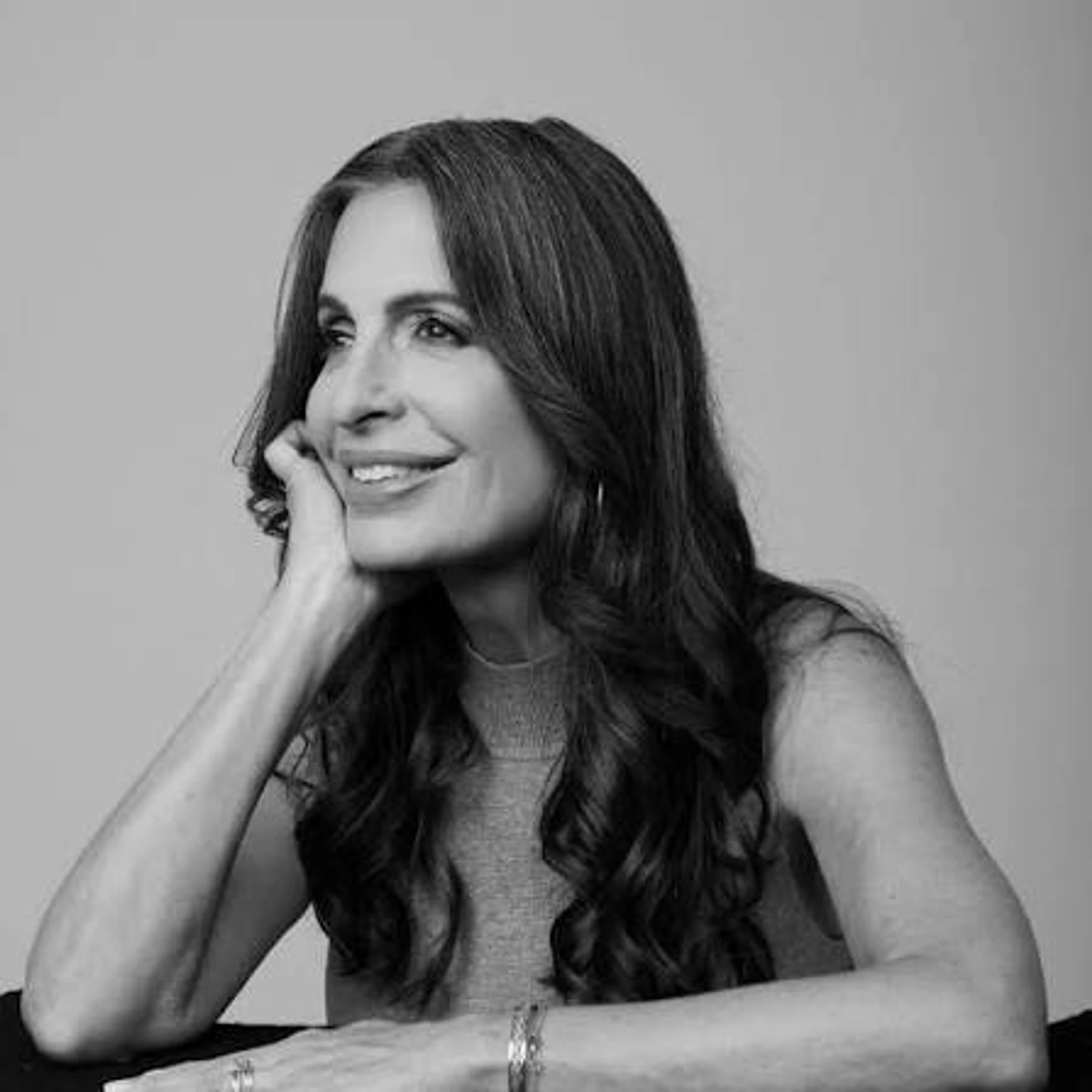 A black and white studio portrait of a smiling woman with long, dark, wavy hair. She rests her chin on her hand, looking thoughtfully to the side.