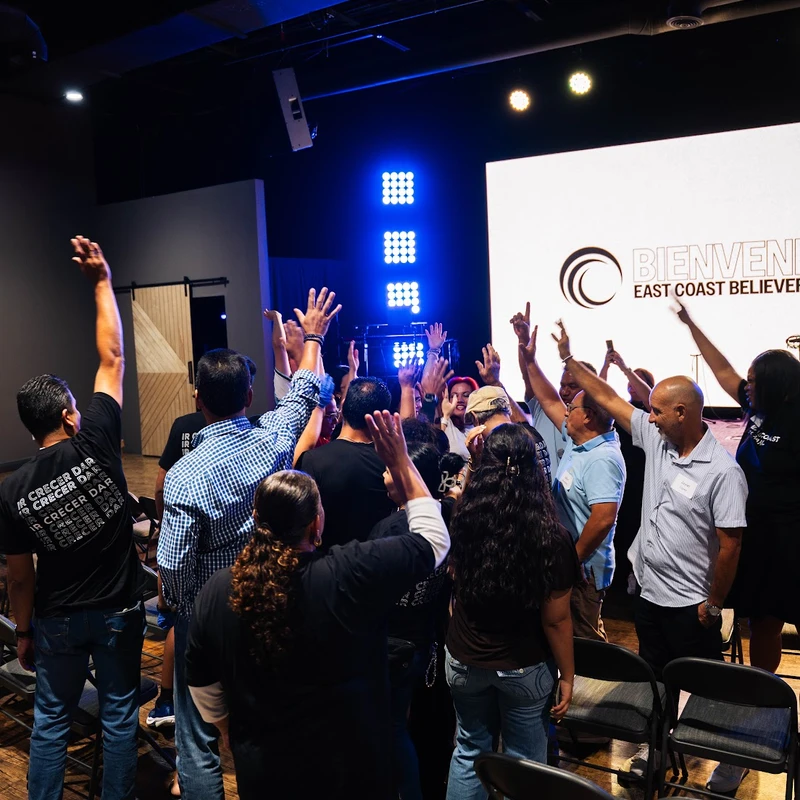 A group of people, seen from behind, stand with their hands raised towards a stage with a screen that reads 'Bienvenidos East Coast Believers'.