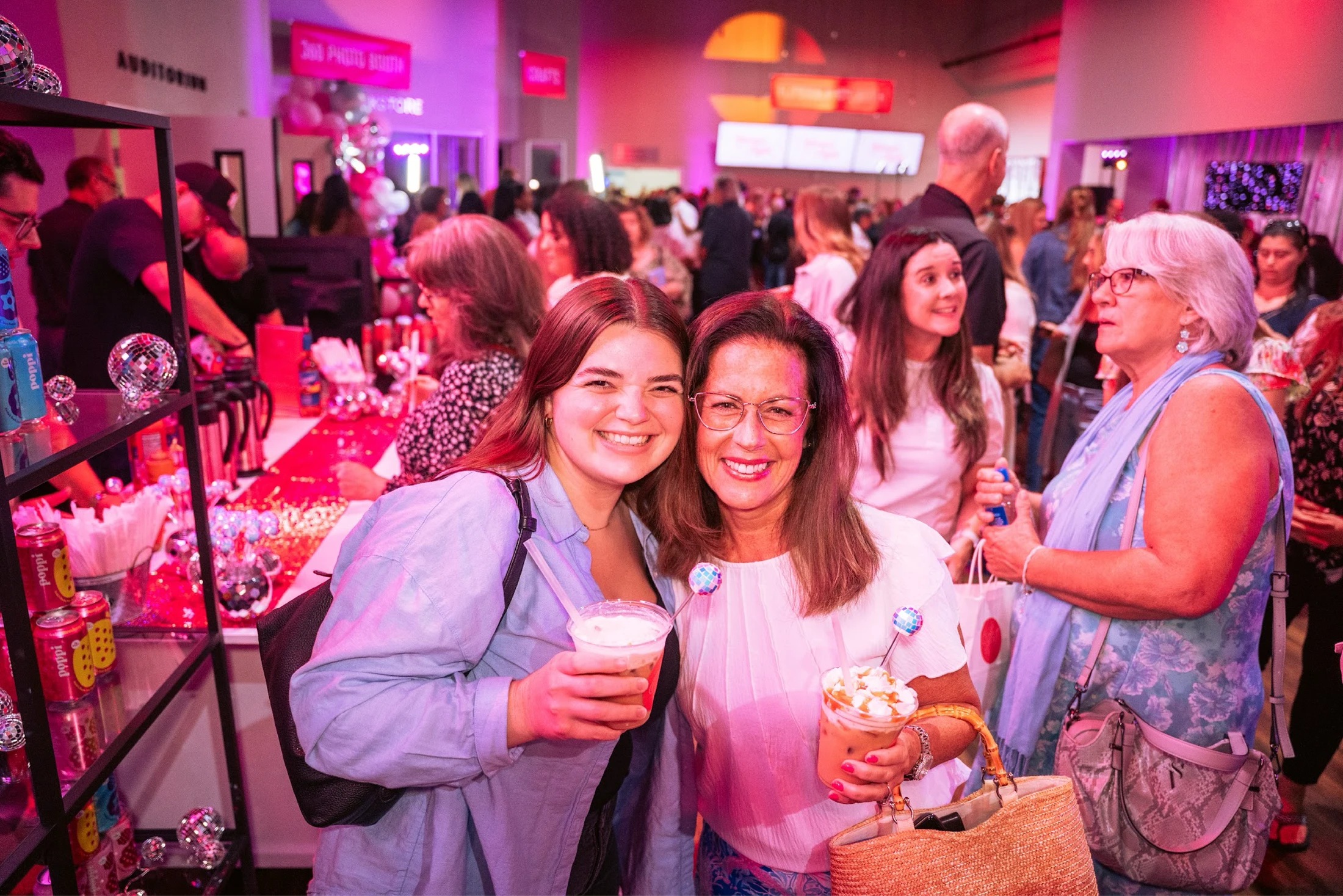 Two women smile for the camera while holding iced drinks at a crowded party. The venue is decorated with pink lights, balloons, and small disco balls.