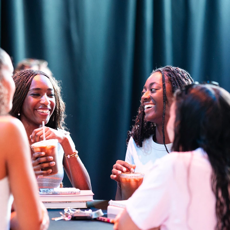 A group of four young women sit at a table, talking and laughing. Two Black women in the center smile at each other, one holding an iced coffee.