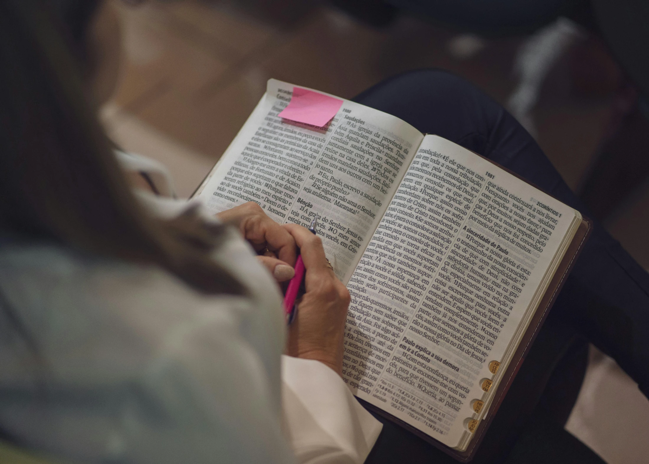 An over-the-shoulder view of a woman sitting with an open Bible on her lap, holding a pink pen over the text as she studies.