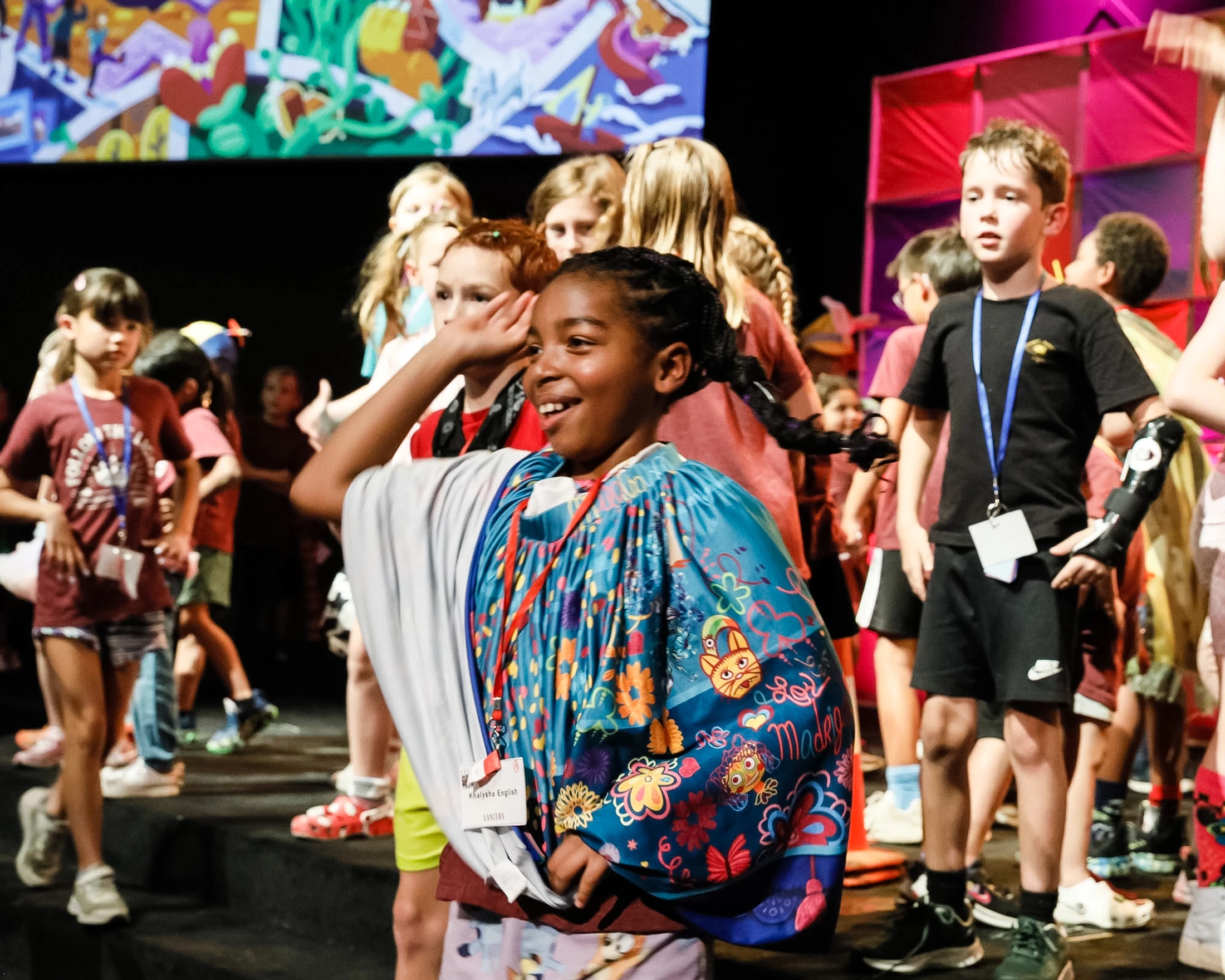 A young Black girl with braided hair smiles on stage, wearing a colorful blue cape. Other children stand around her under bright stage lights.