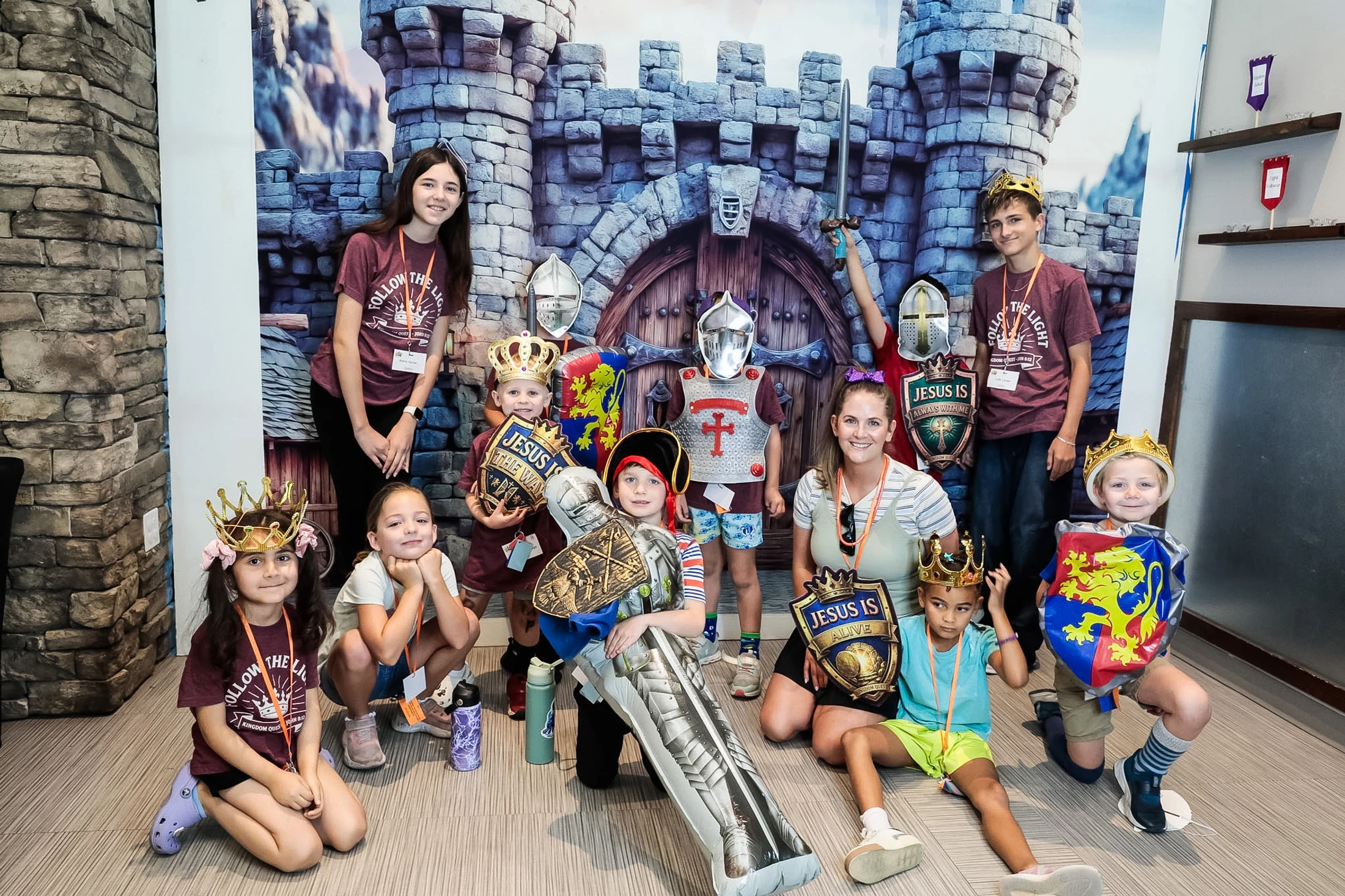 A group of children and a woman pose in medieval costumes with crowns, helmets, and shields in front of a castle backdrop for a Vacation Bible School event.