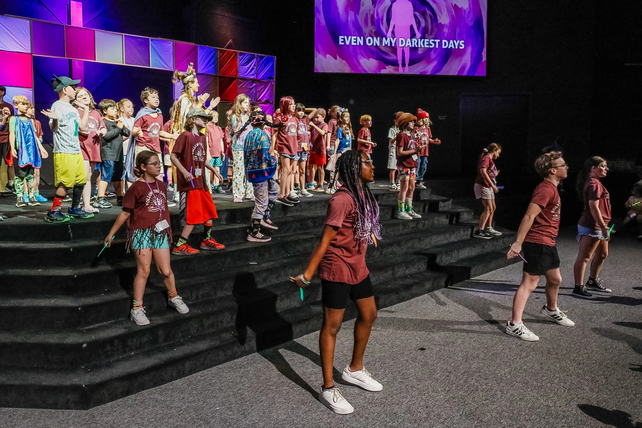 A large group of children in matching maroon t-shirts perform on a tiered stage under purple lights, with a screen displaying song lyrics behind them.
