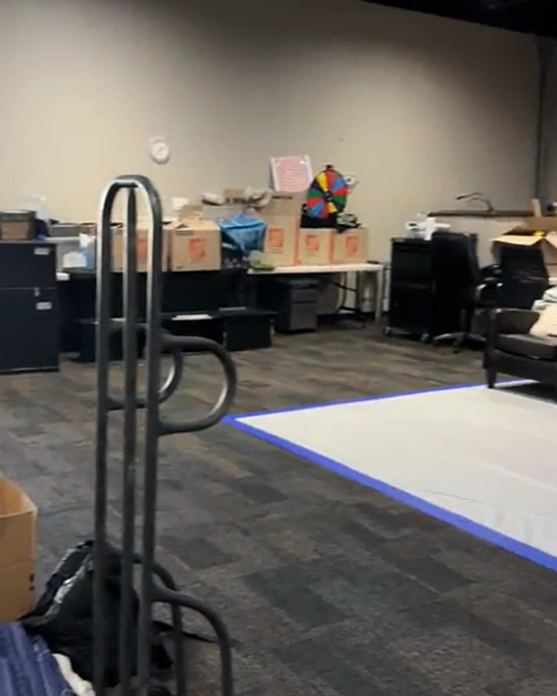 A cluttered office space with moving boxes on a table, office chairs, a hand dolly in the foreground, and a white rectangle taped to the carpeted floor.