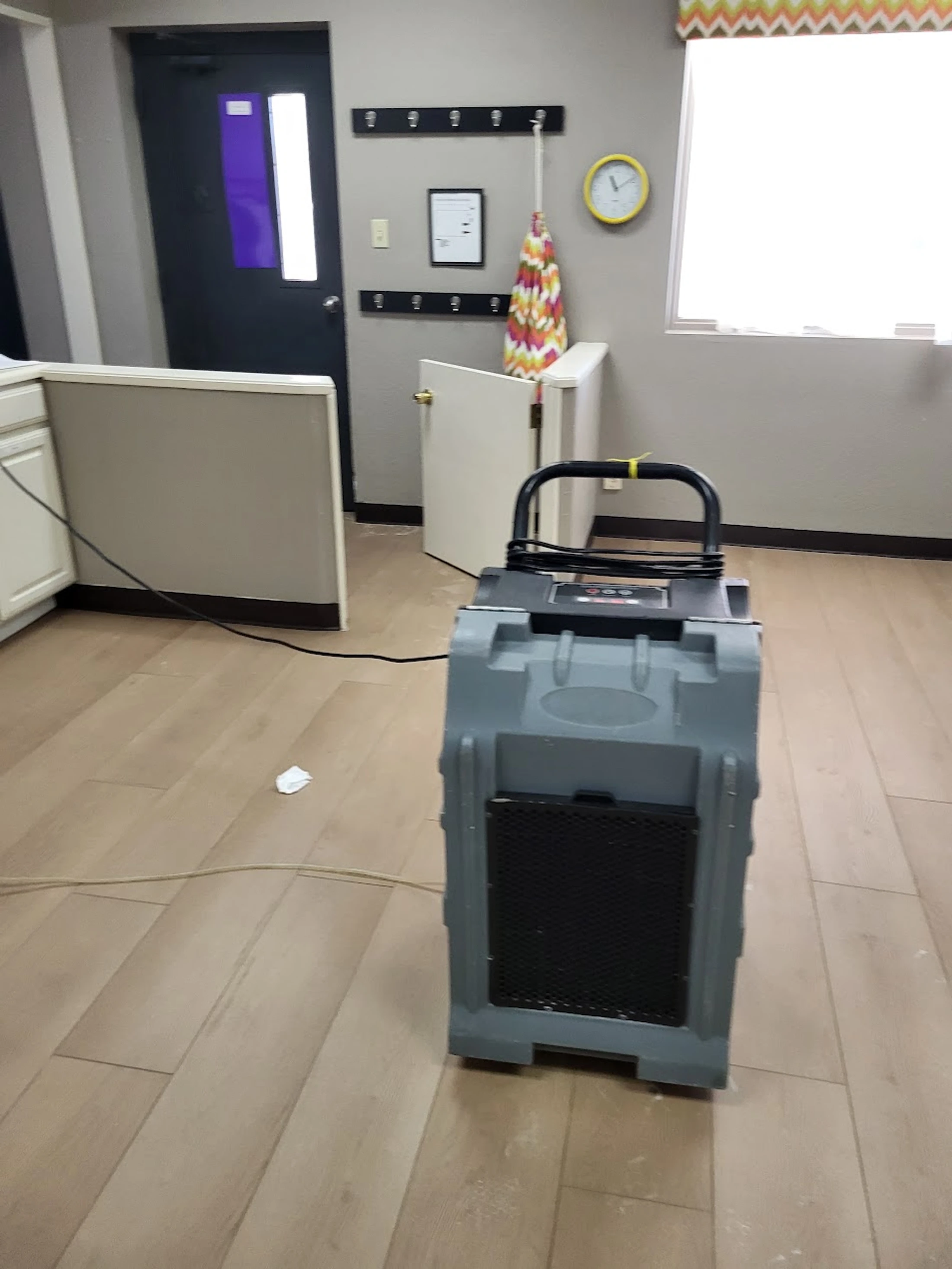 A large, grey industrial dehumidifier sits on a light wood-look floor in an office setting, likely being used for water damage restoration.