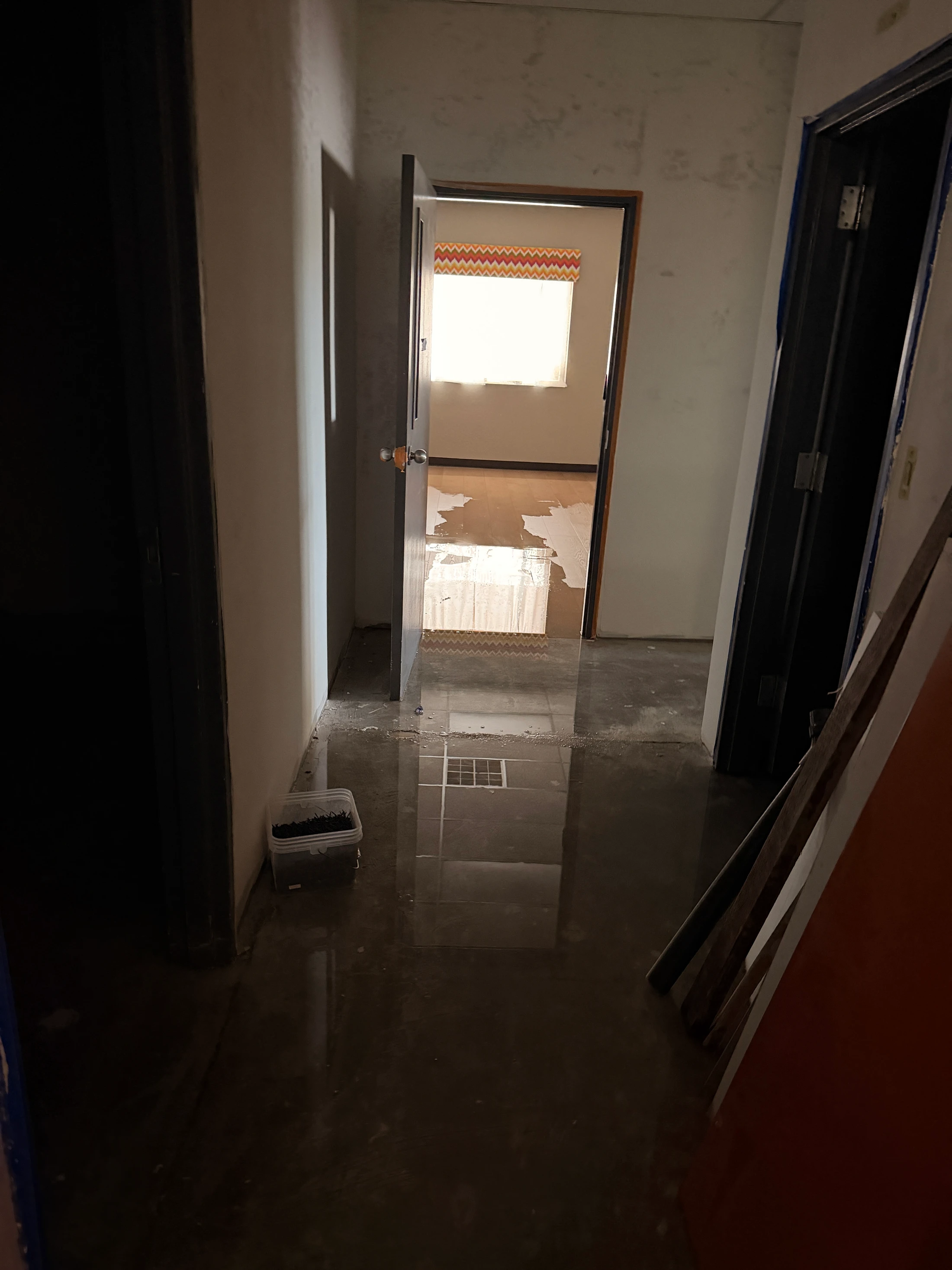 A dark hallway with a flooded, reflective floor during a home renovation. An open door reveals a brighter room with a window and partially finished flooring.