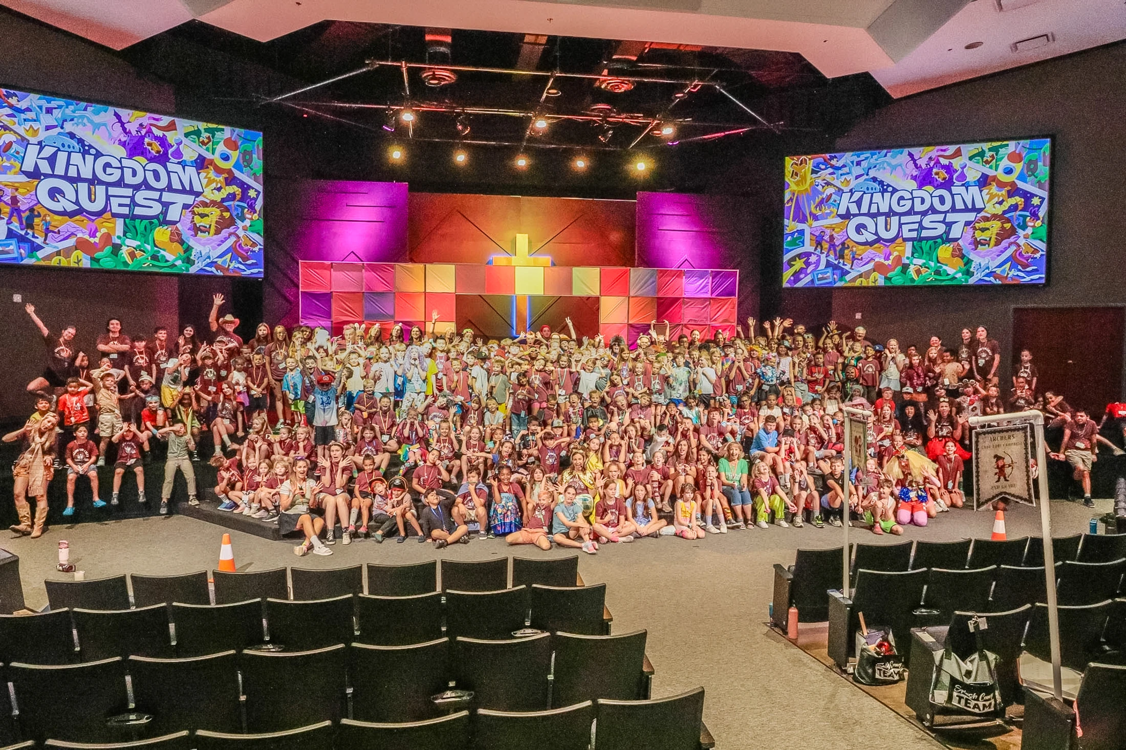 A large group of children and adults posing for a group photo on a colorful stage under two screens that read 'Kingdom Quest'.