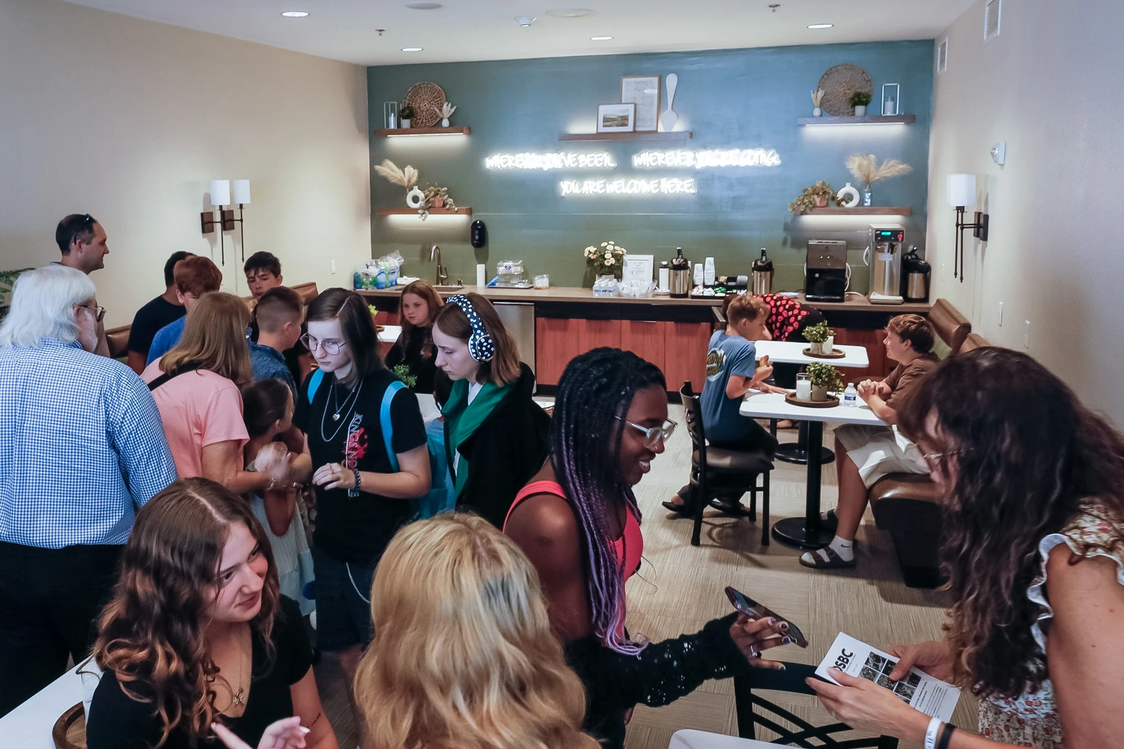 A diverse group of people socializes in a modern room with a coffee bar and a neon sign on a blue wall that says 'You are welcome here.'