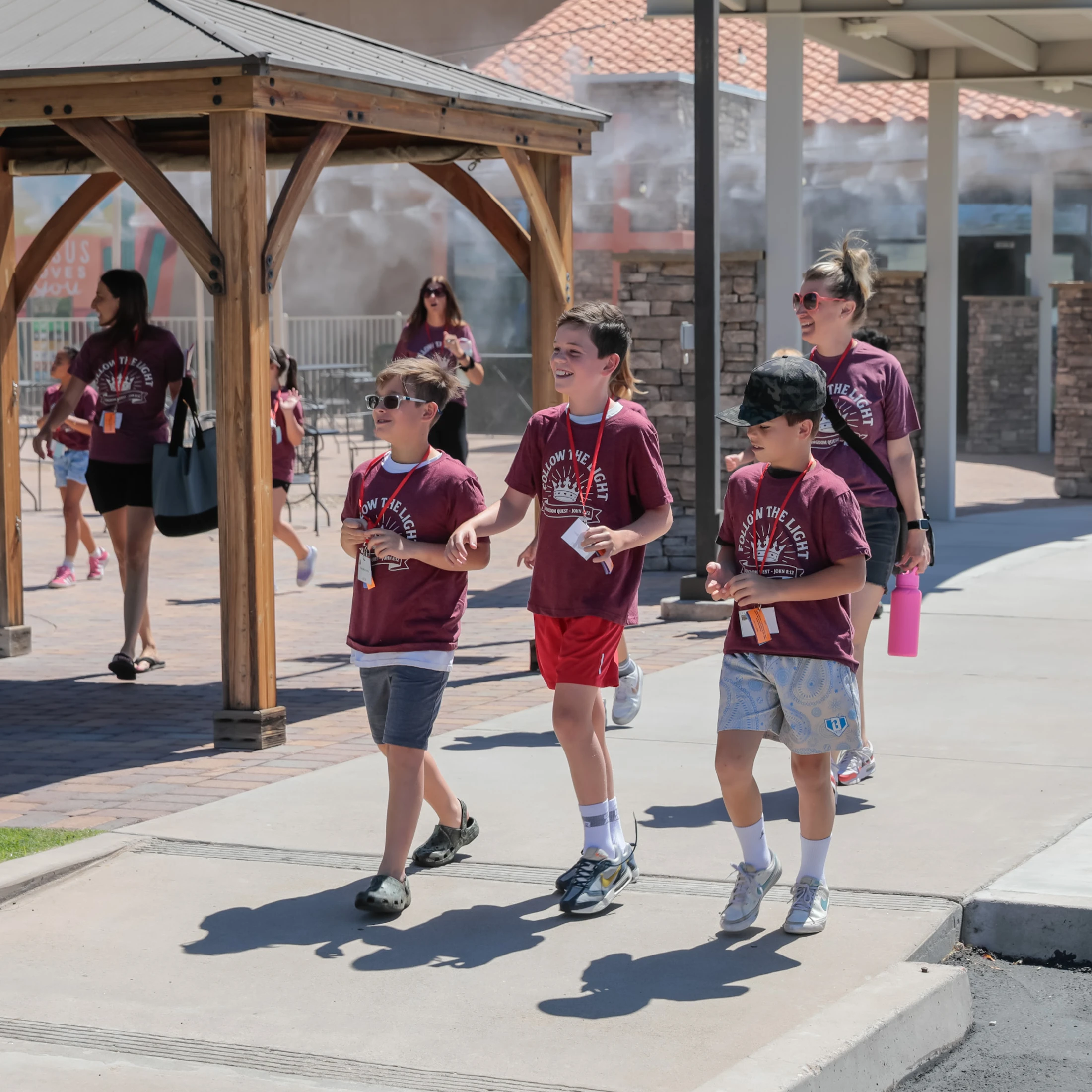 A group of three boys in matching maroon t-shirts walk and smile on a sunny sidewalk, followed by adults. Misters spray a cool mist in the background.