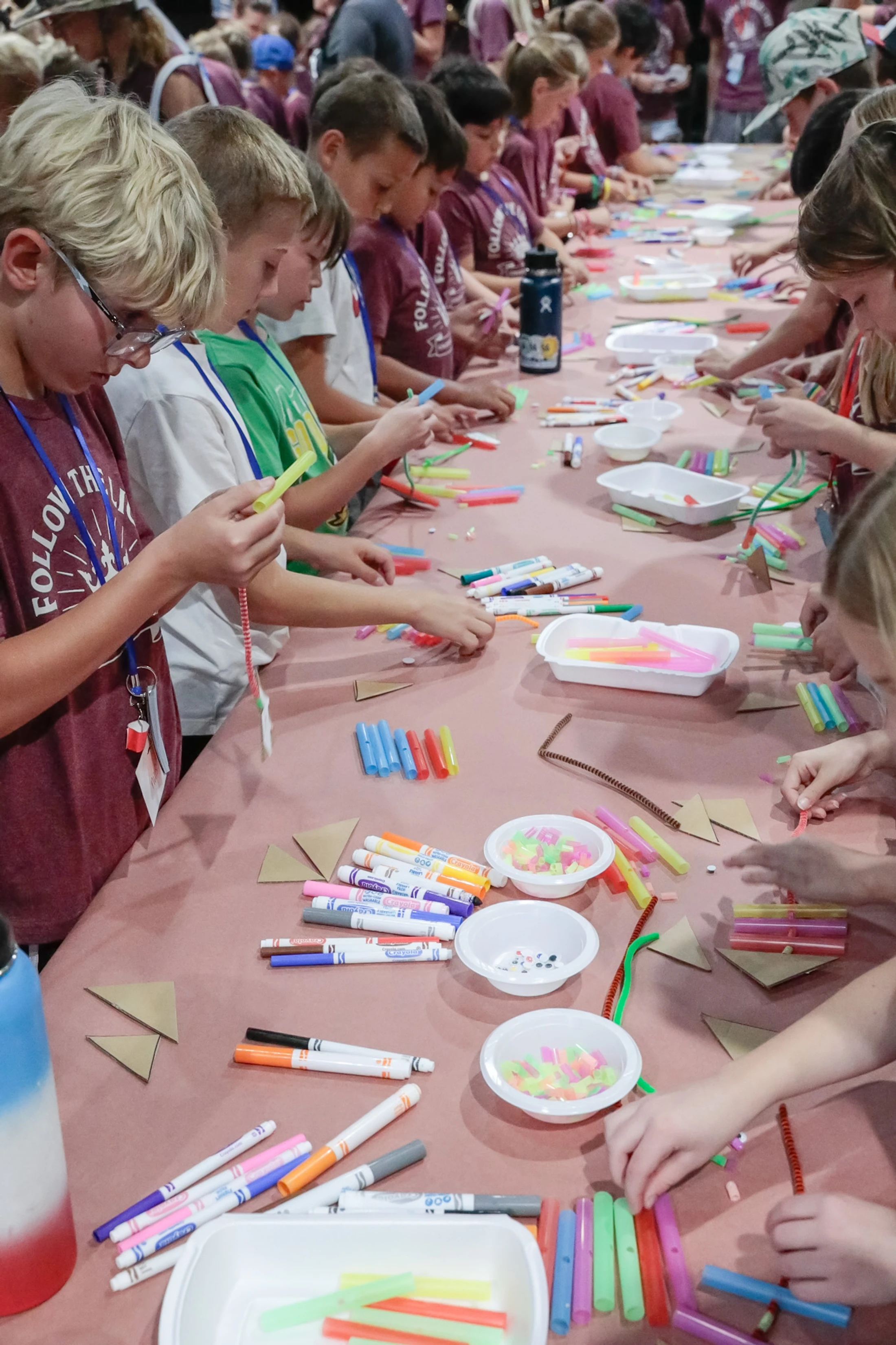A long table lined with children focused on making crafts with colorful supplies like markers, straws, and pipe cleaners at a group event.