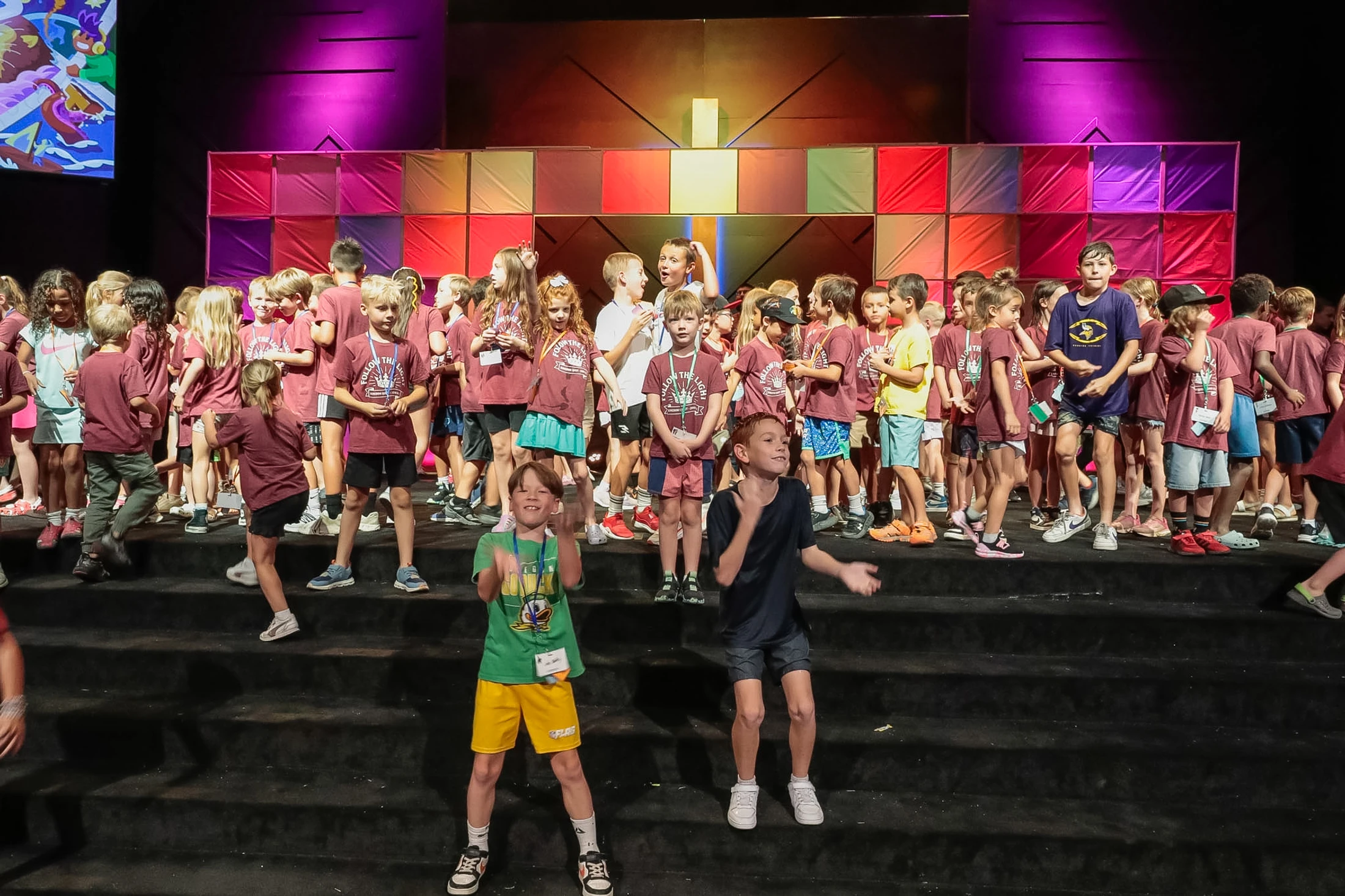 A large group of children stand on a stage with a colorful block background, singing. In the foreground, two boys dance enthusiastically on the steps.