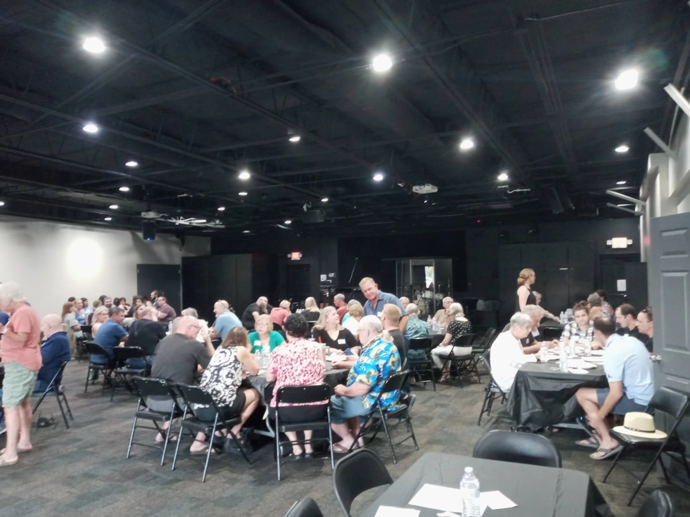 A wide shot of a large, dimly lit event space with a black ceiling, filled with people sitting at round tables and socializing with each other.