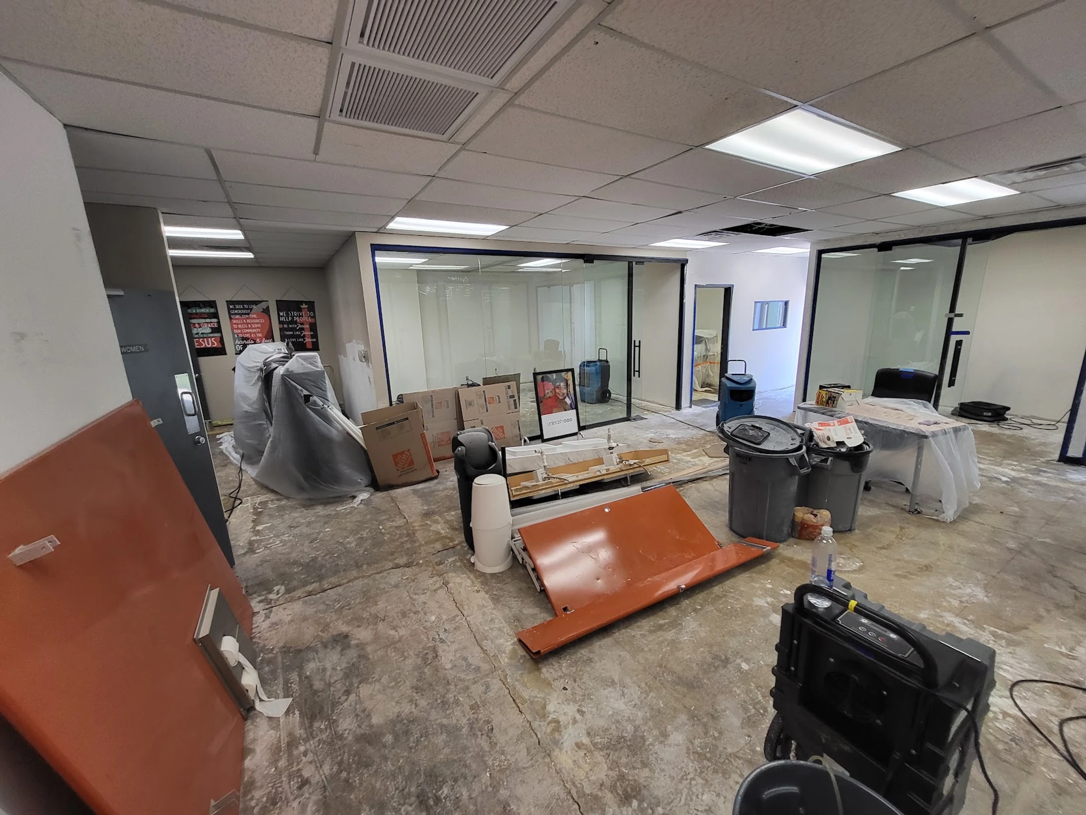 An office interior in disarray during renovation, with bare concrete floors, scattered boxes, covered furniture, and industrial air movers.