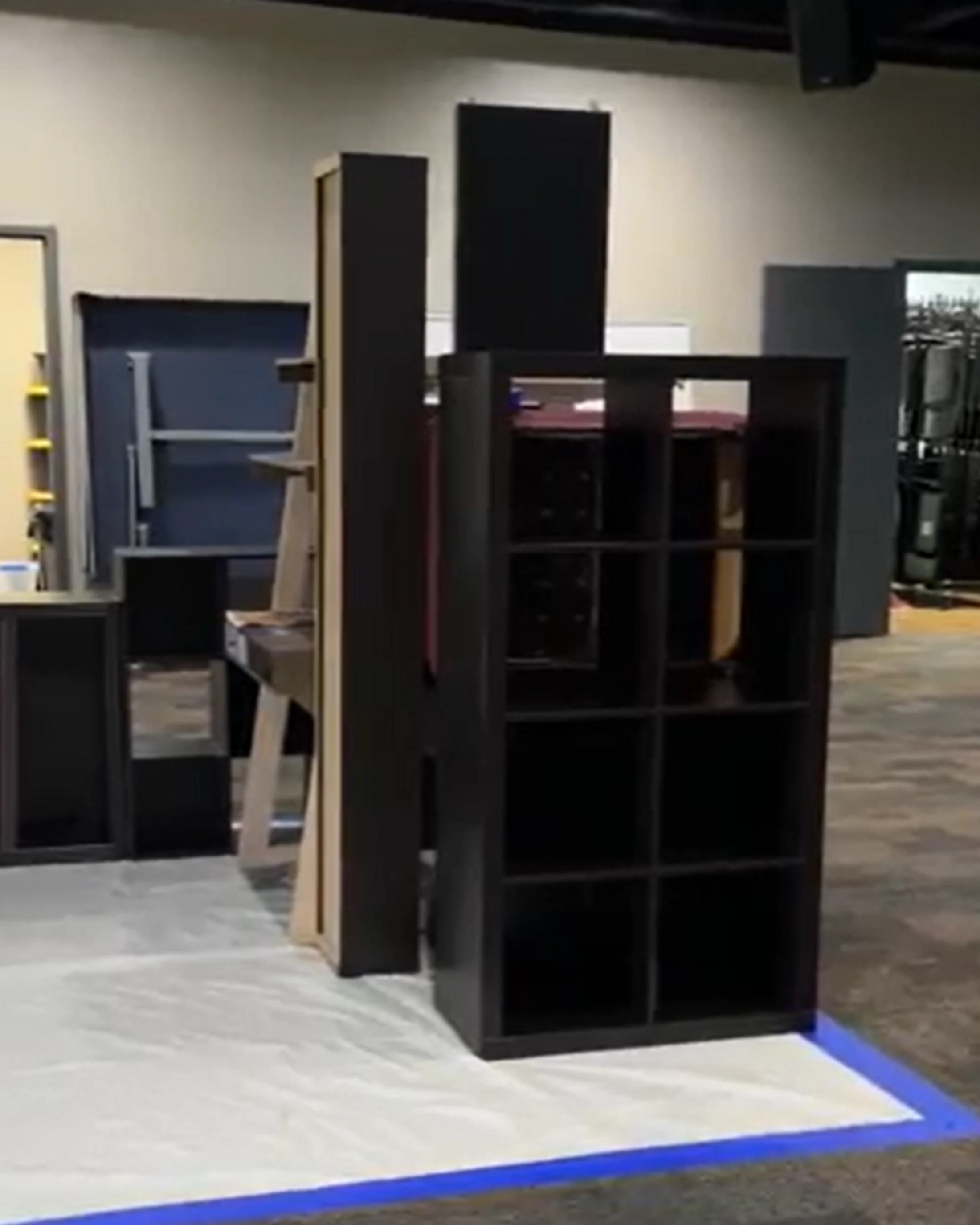 A dark brown, eight-cubby shelving unit stands on a white drop cloth taped to the floor, with other furniture and sound panels in the background.