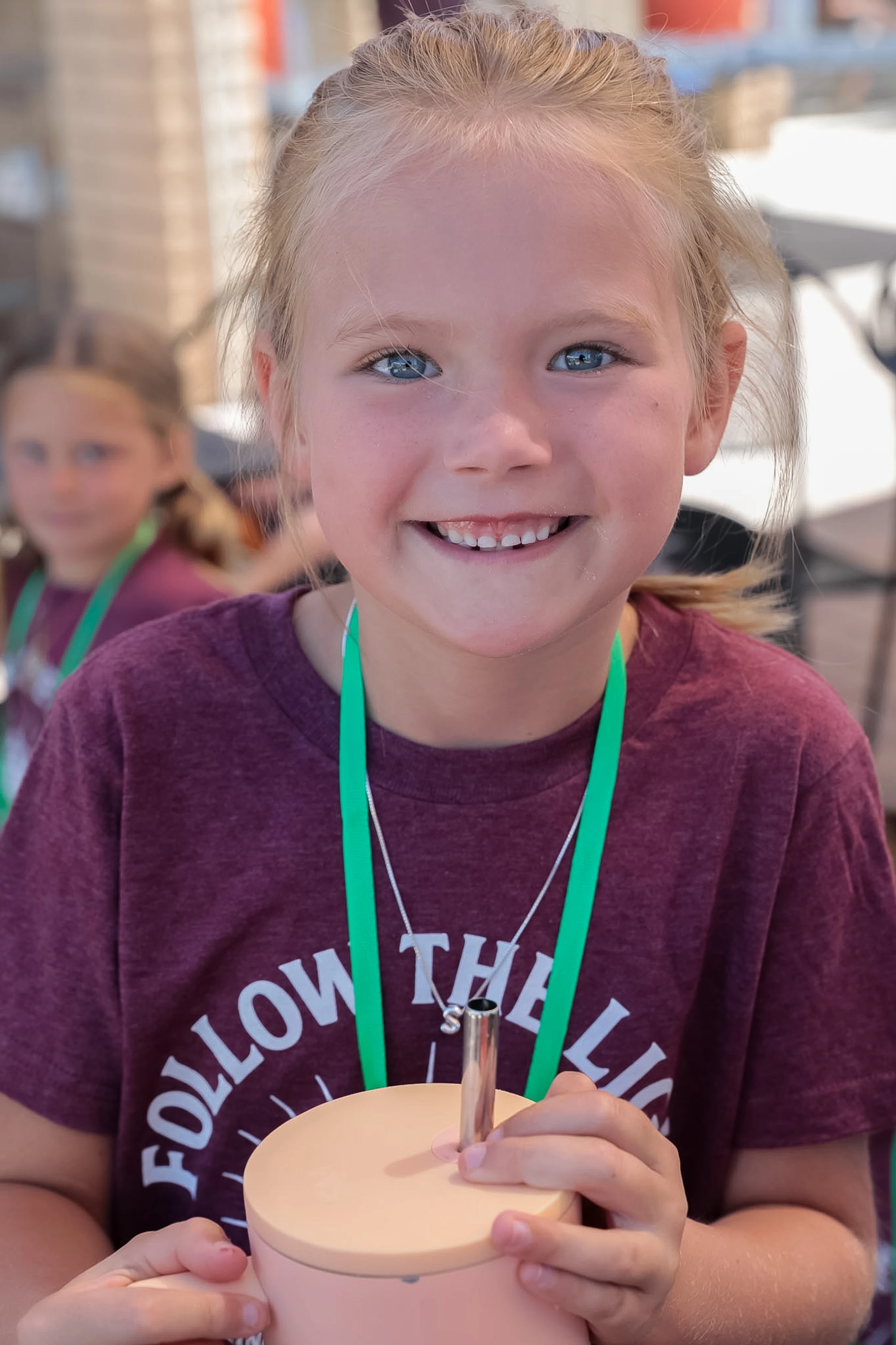 A young blonde girl with blue eyes smiles at the camera. She wears a maroon shirt, a green lanyard, and holds a pink tumbler with a metal straw.