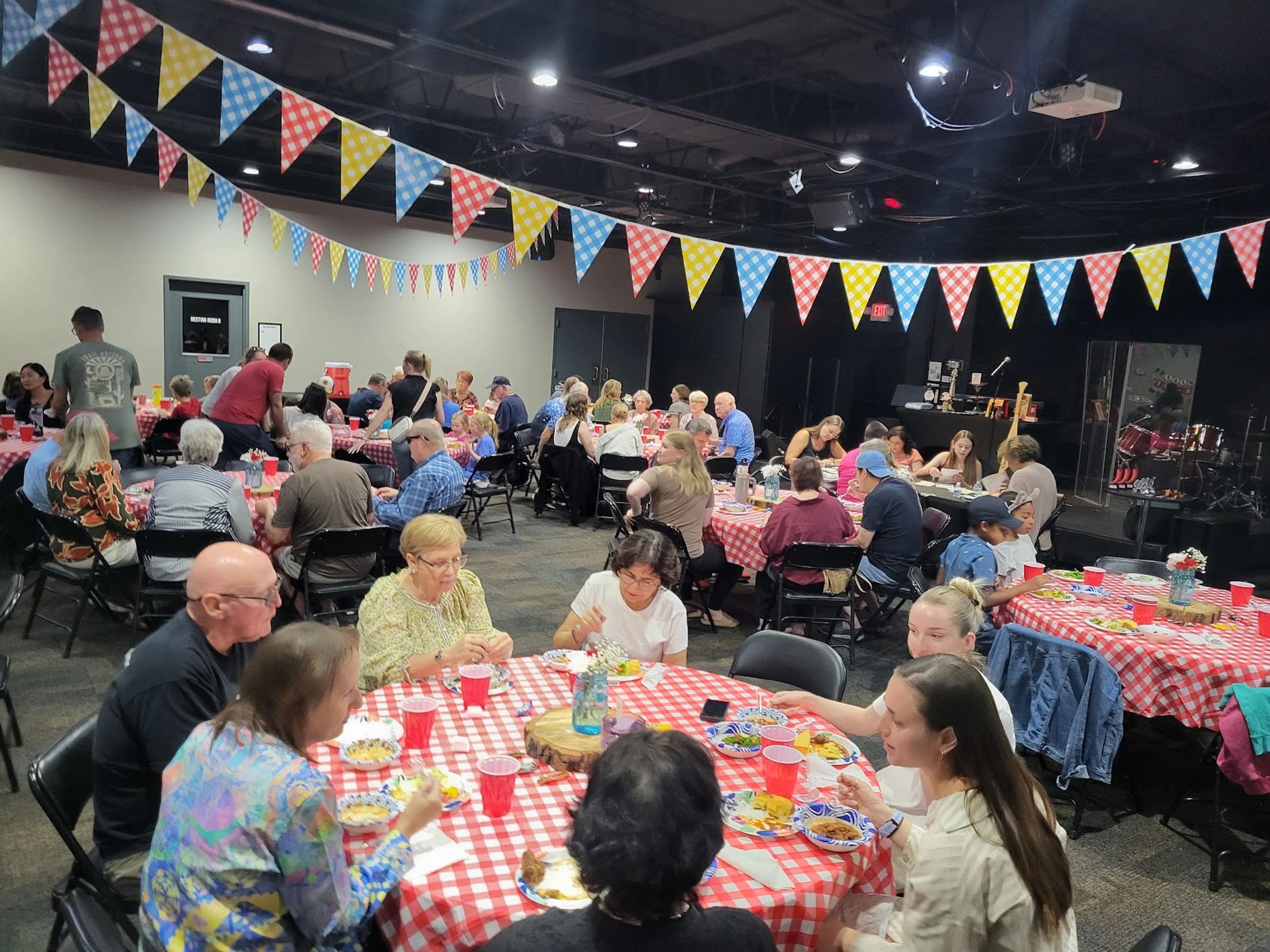 A large group of people sits at round tables with red checkered tablecloths, enjoying a meal together in a hall decorated with colorful pennant banners.