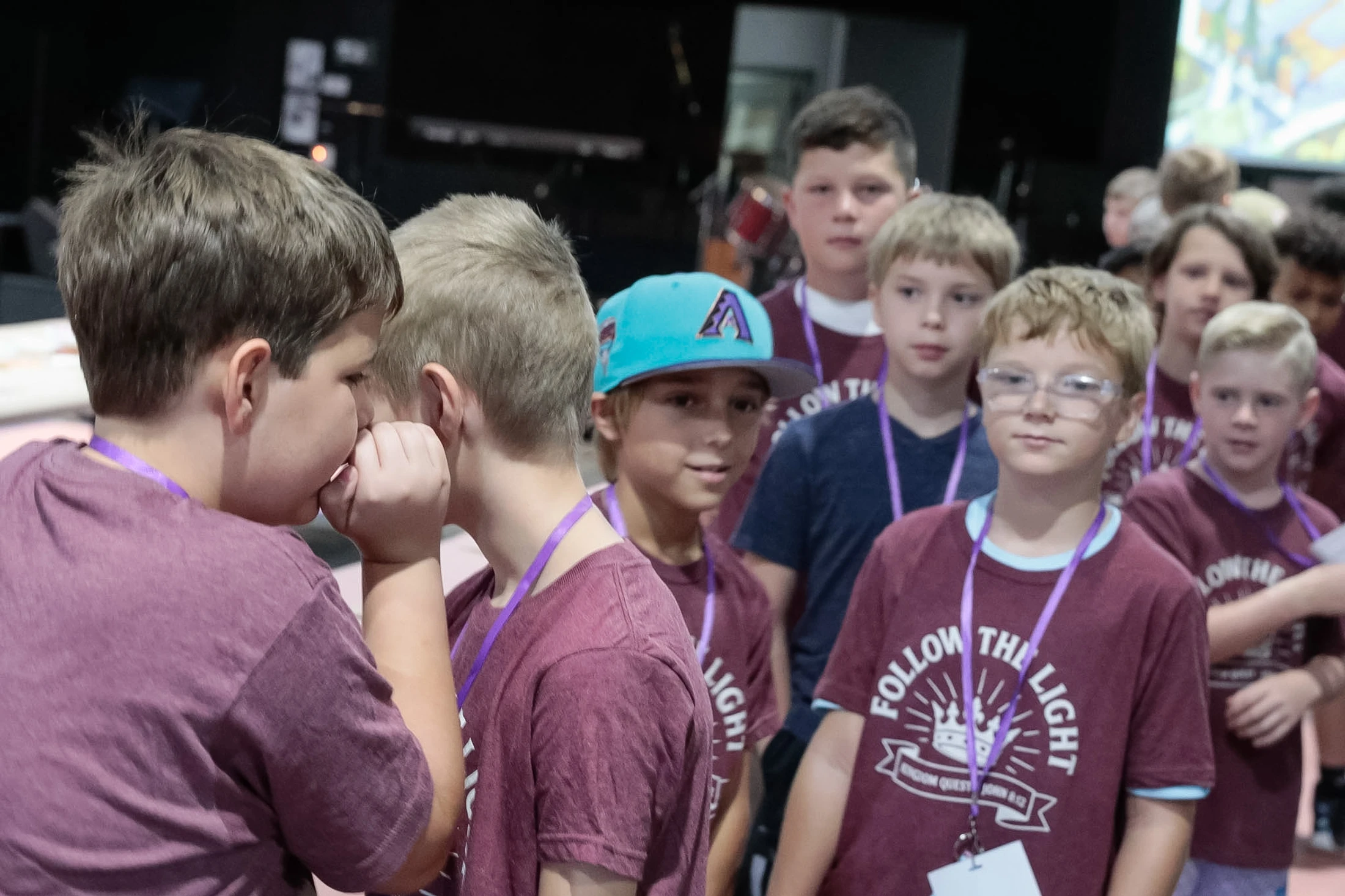 A group of young boys in matching maroon shirts stand in a line. In the foreground, one boy cups his hands around his mouth to whisper into another's ear.
