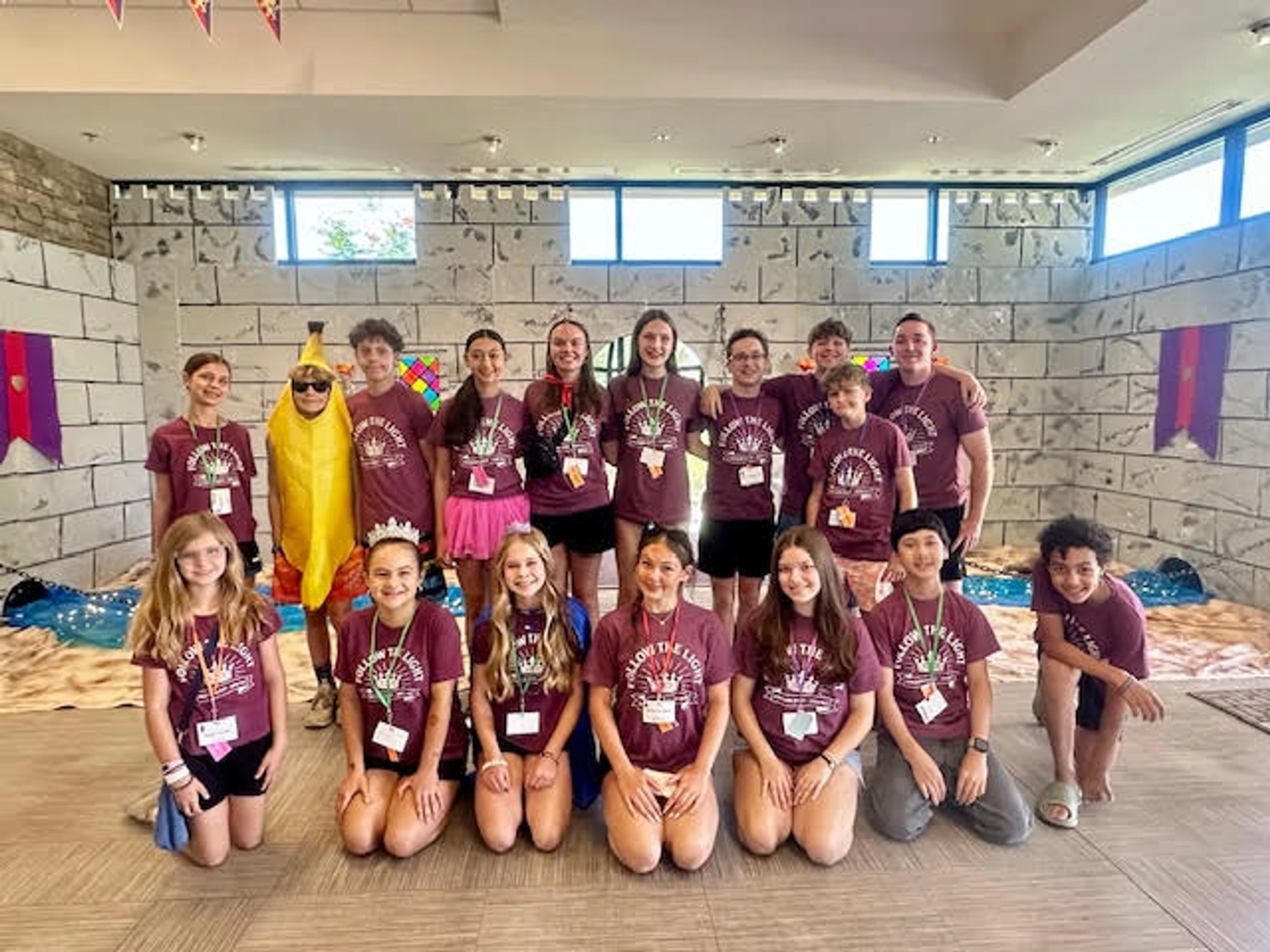 A large group of smiling kids and teens pose for a photo. Most wear matching maroon t-shirts, one wears a banana costume, and the room looks like a castle.