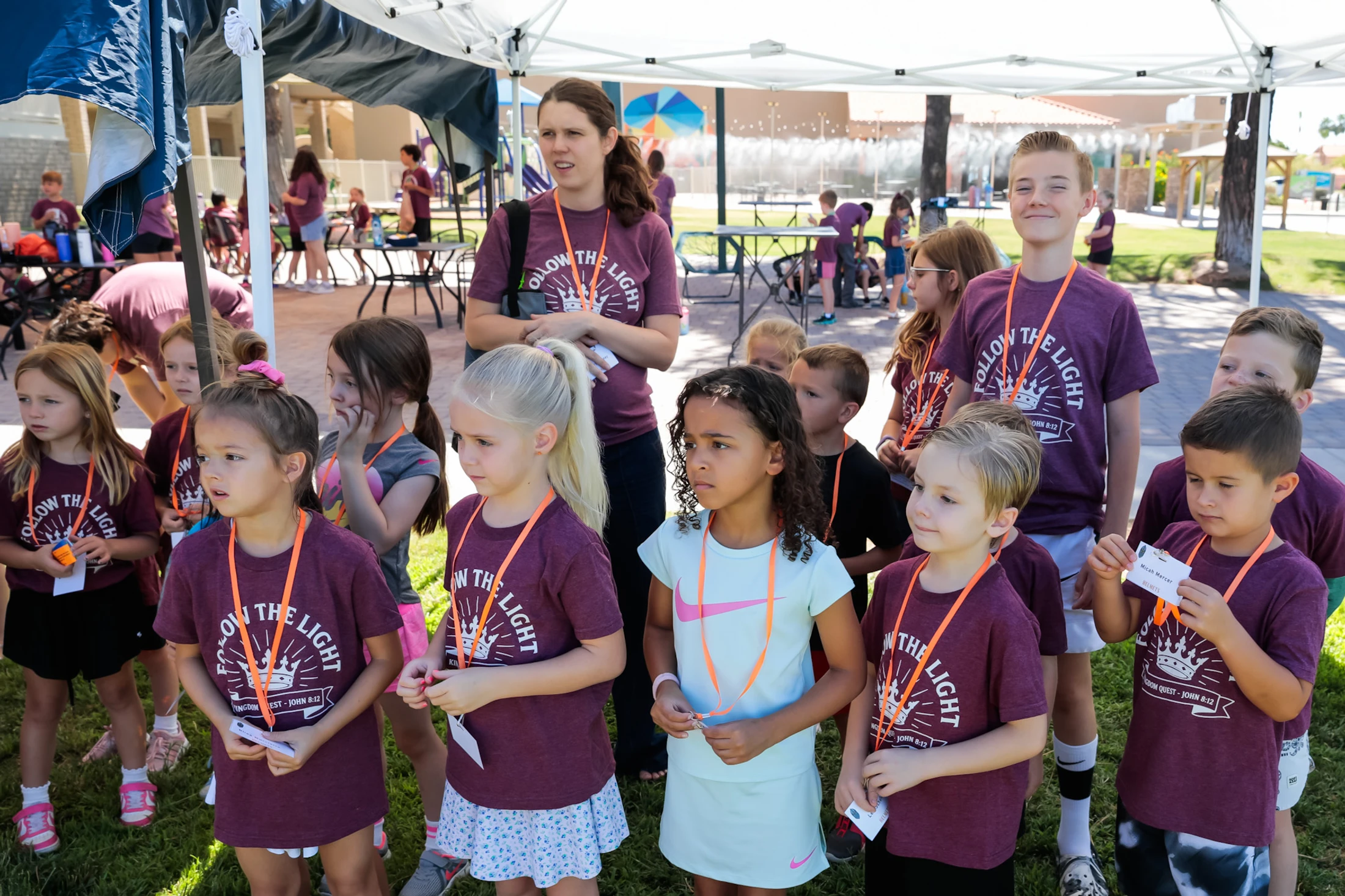 A group of elementary-aged children and a female leader stand under an outdoor tent, wearing matching maroon shirts with 'Follow the Light' printed on them.