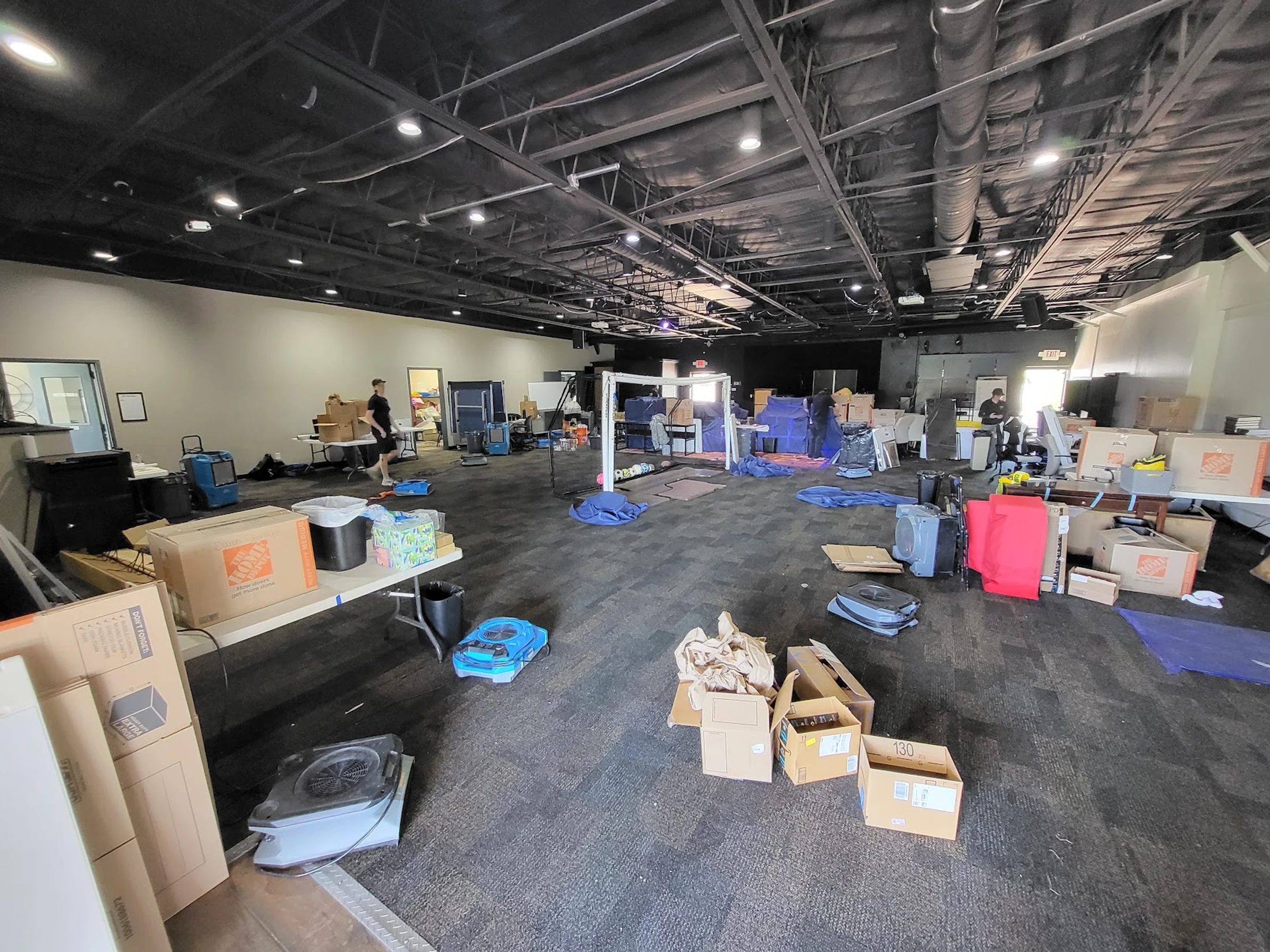 A wide shot of a large, open-plan room with a dark carpet and industrial ceiling, filled with moving boxes, tables, and equipment in a state of disarray.