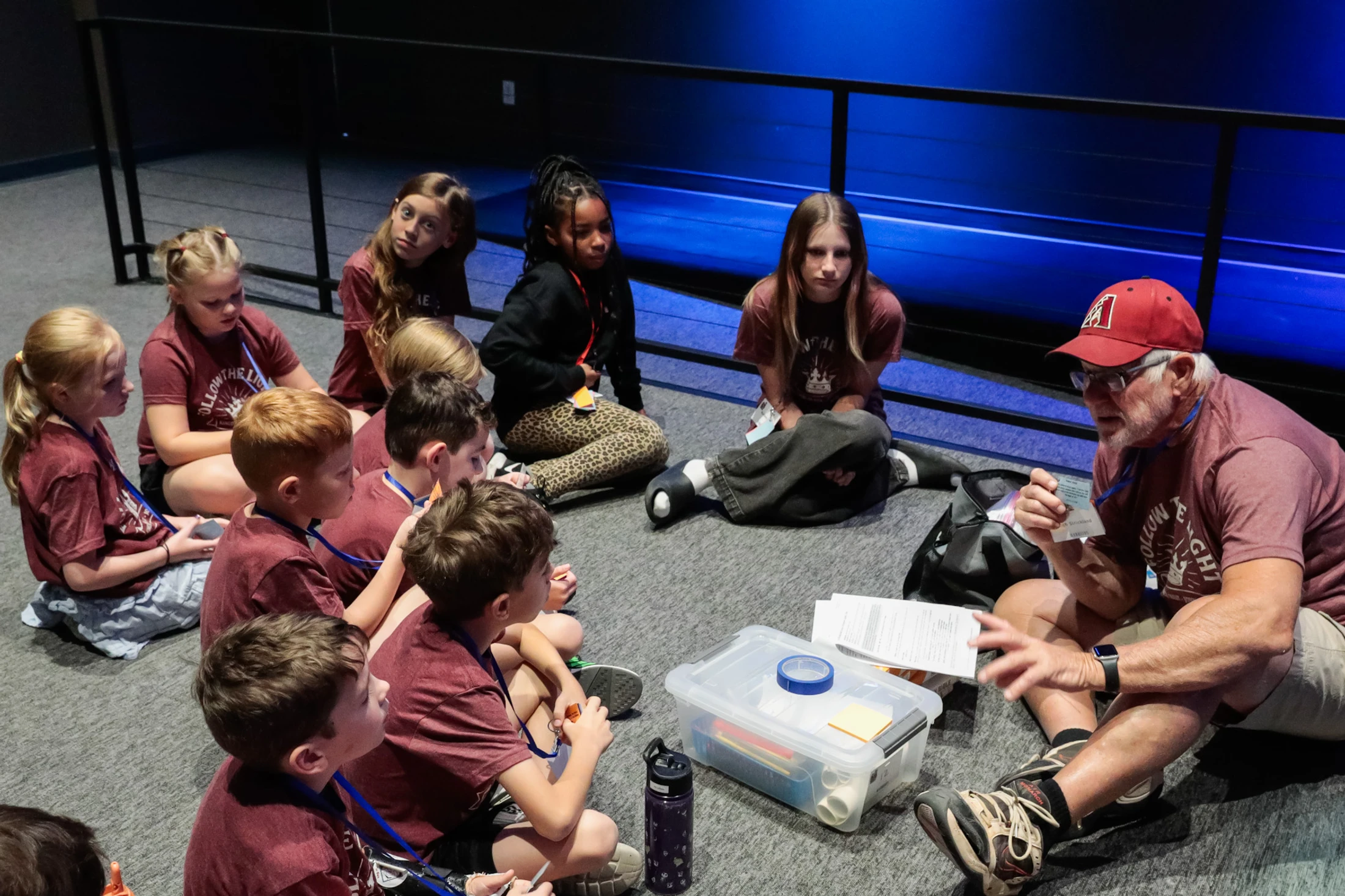 An older man in a red cap sits on the floor with a group of diverse children, teaching them a lesson as they listen attentively in a circle.
