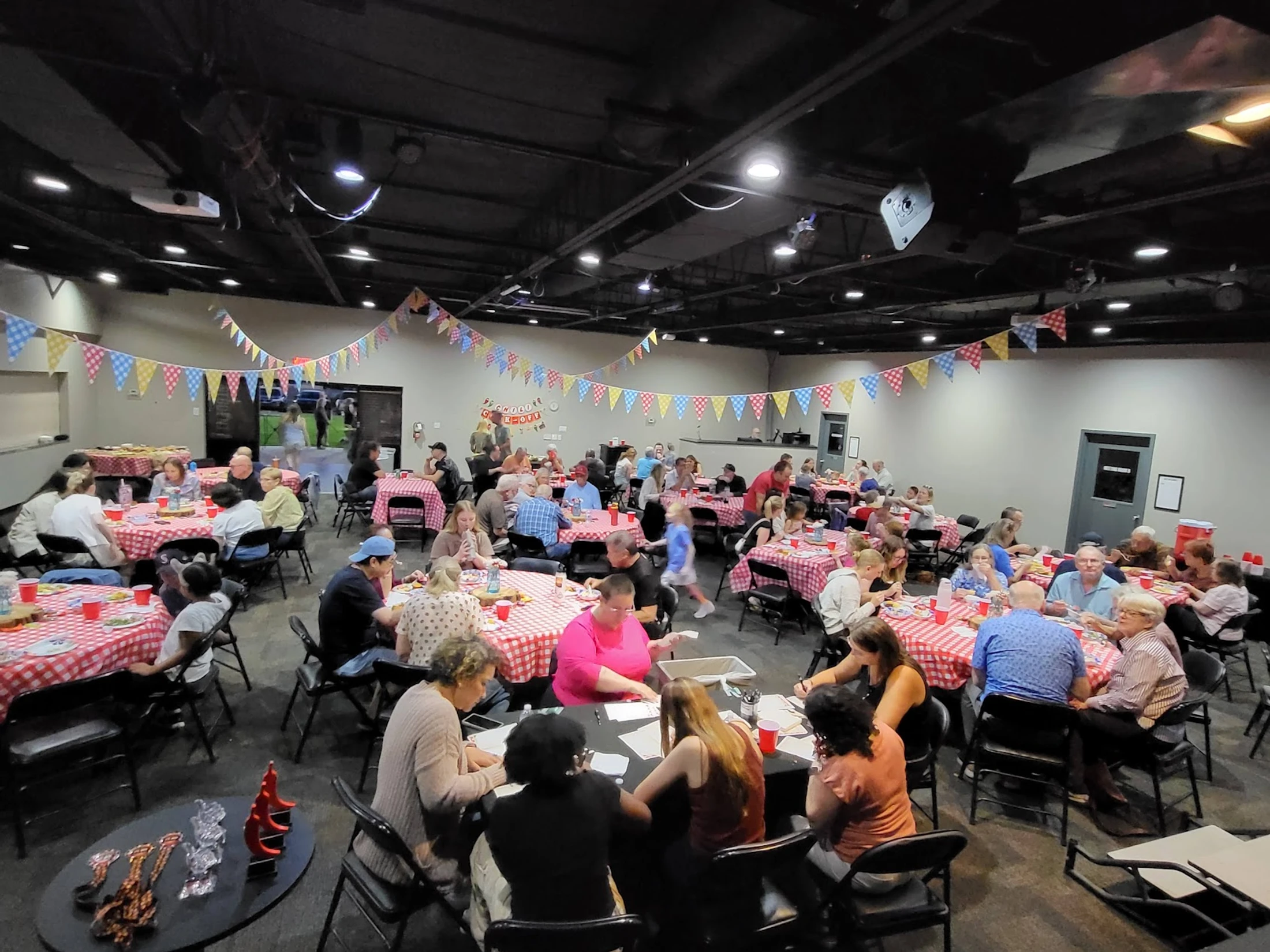 A large hall filled with people socializing and eating at tables with red and white checkered tablecloths. Colorful pennant banners hang from the ceiling.