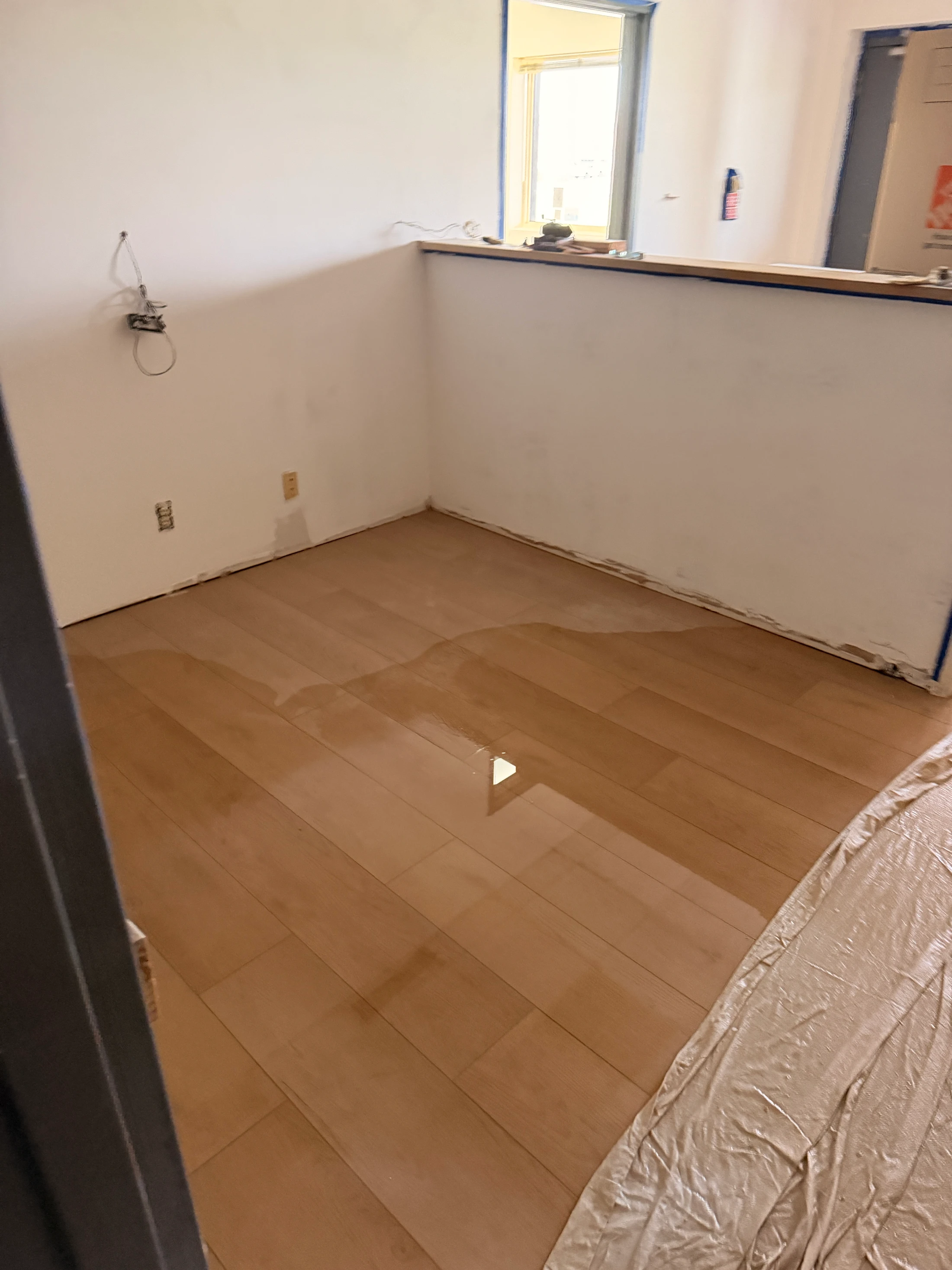 A large puddle of water sits on a light-colored wood floor in the corner of a room that is being renovated, with white, unfinished walls.