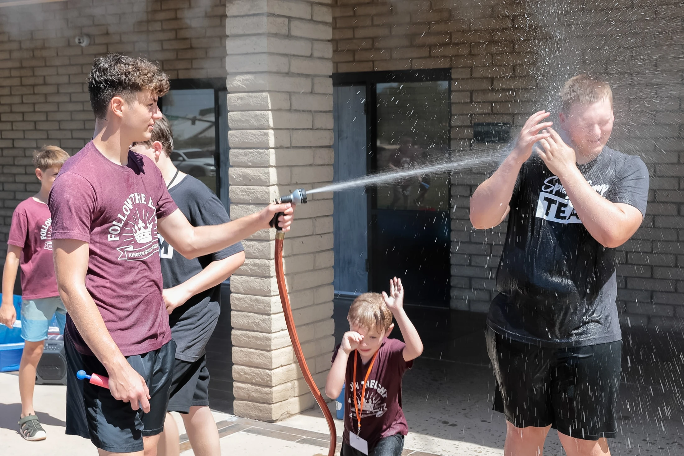 A teenager in a maroon shirt sprays another teen in the face with a water hose on a sunny day, while a small child rubs their eyes nearby.