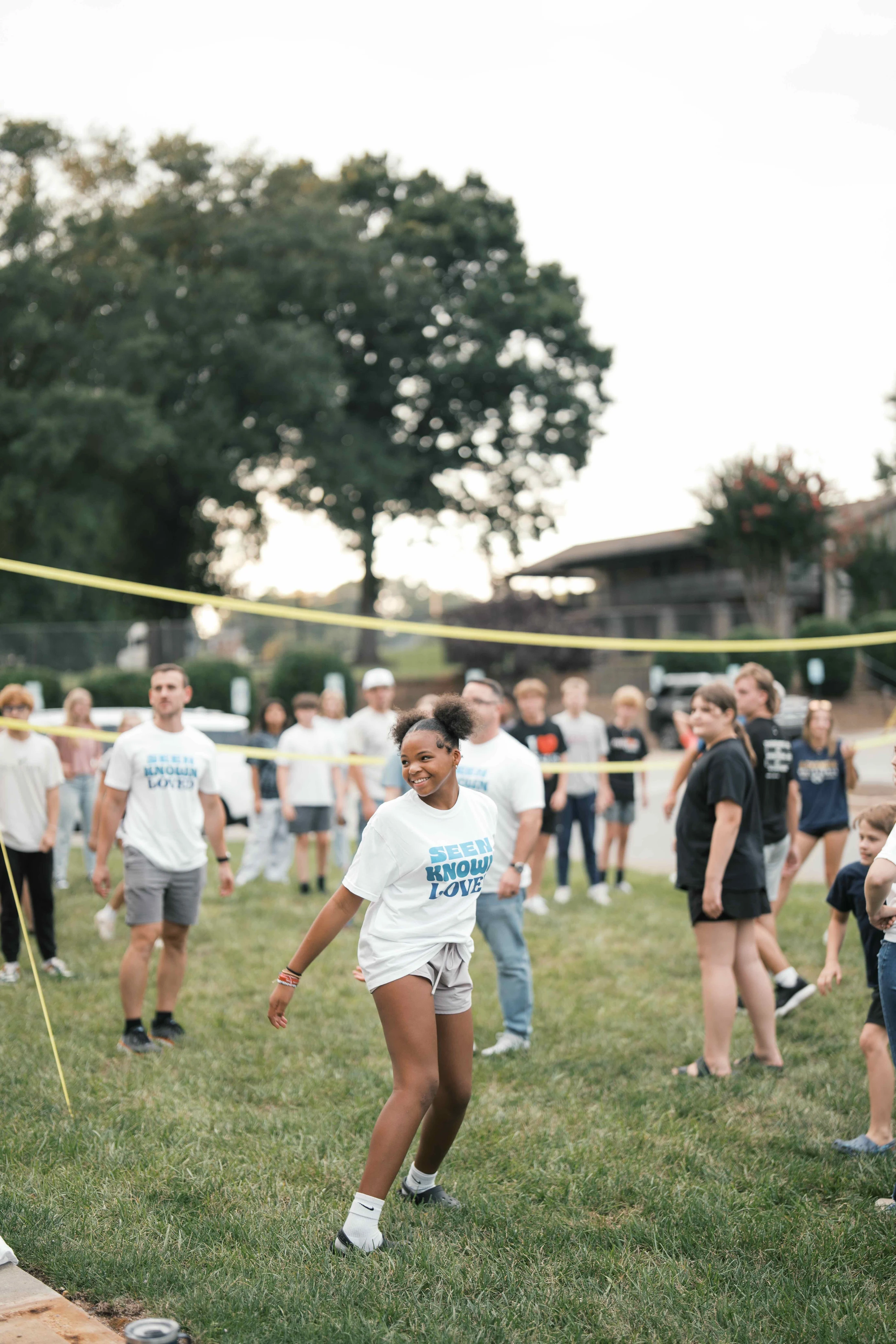 A young Black girl with her hair in two puffs smiles while playing volleyball on a grassy field with a large group of people in the background.
