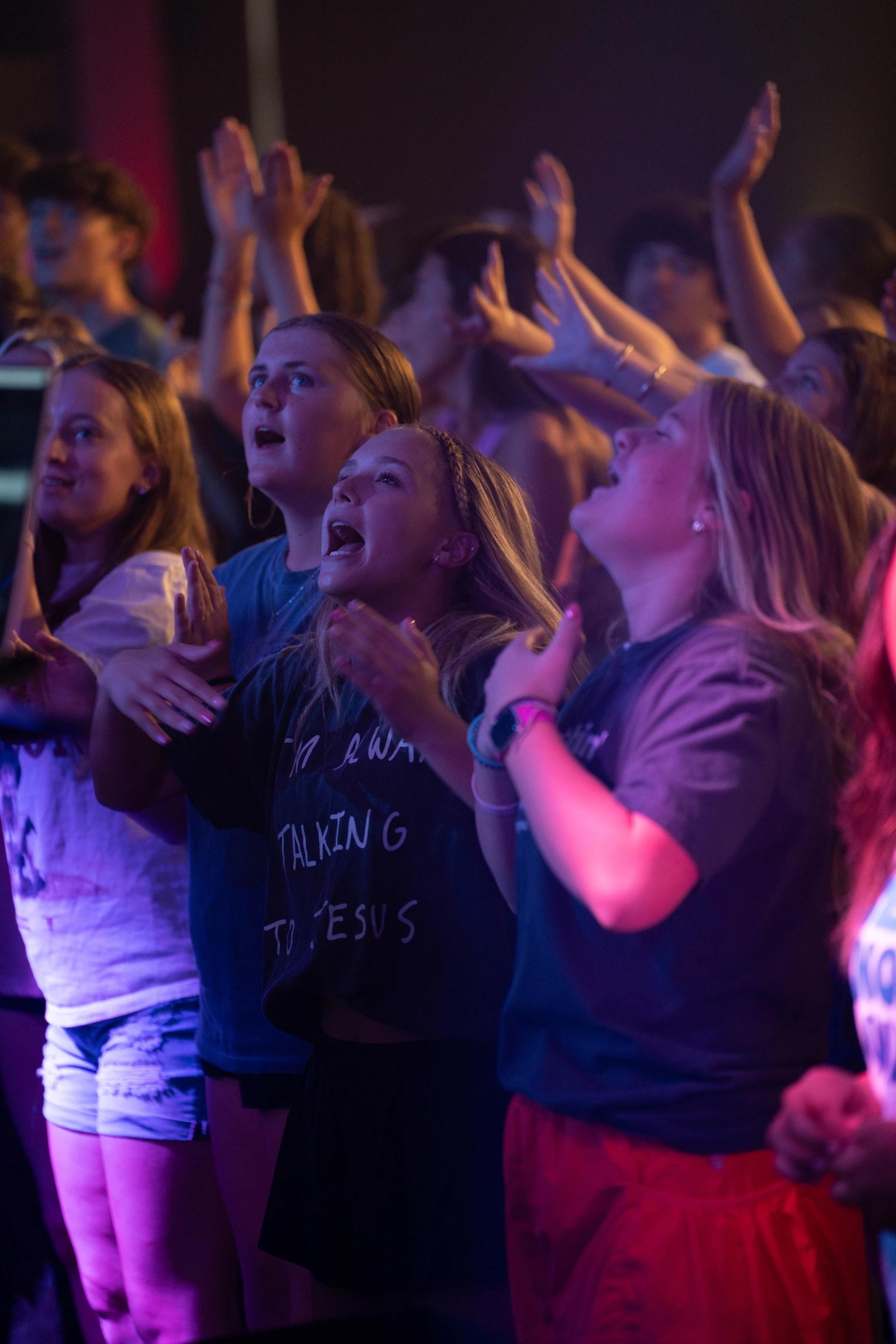 A crowd of young women sing with hands raised in a dimly lit room with purple and pink light, looking up with joyful and worshipful expressions.