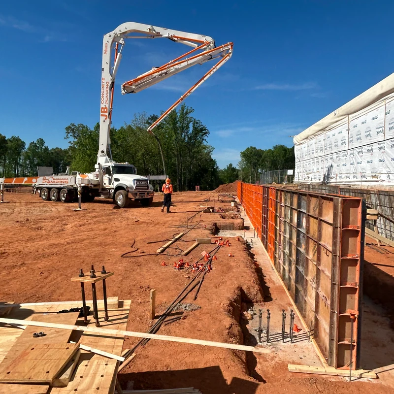 A large concrete pump truck extends its boom arm over foundation forms on a construction site with red dirt under a clear blue sky. A worker looks on.