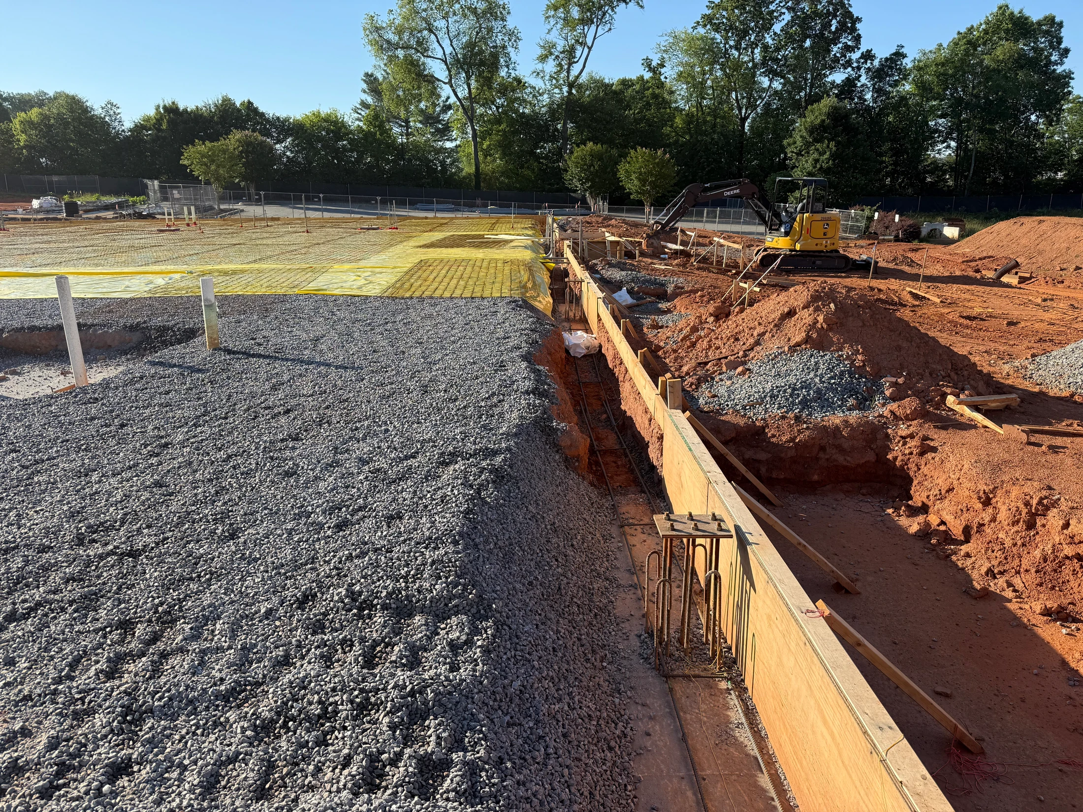 A construction site with foundation work in progress. A gravel base is next to a trench with wooden formwork and rebar, with an excavator in the background.