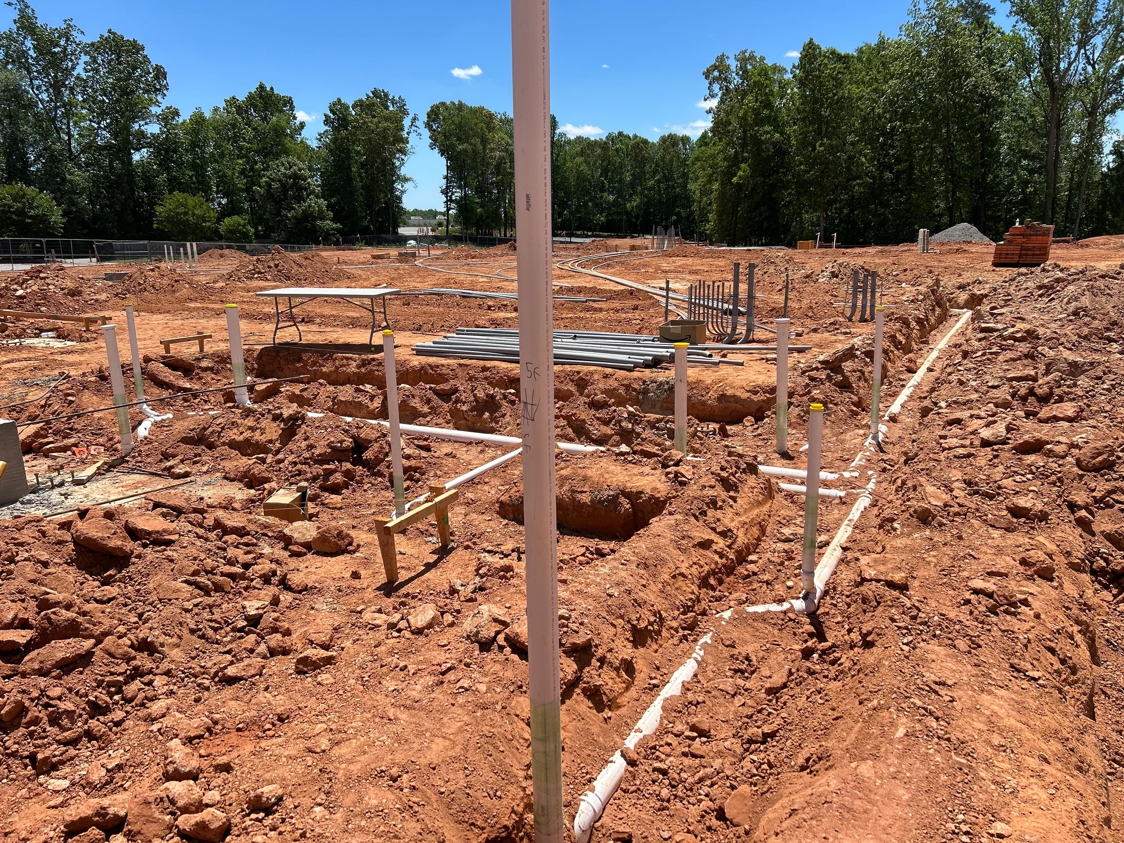 A construction site with red clay soil, where trenches have been dug for the installation of white PVC pipes for the building's foundation plumbing.