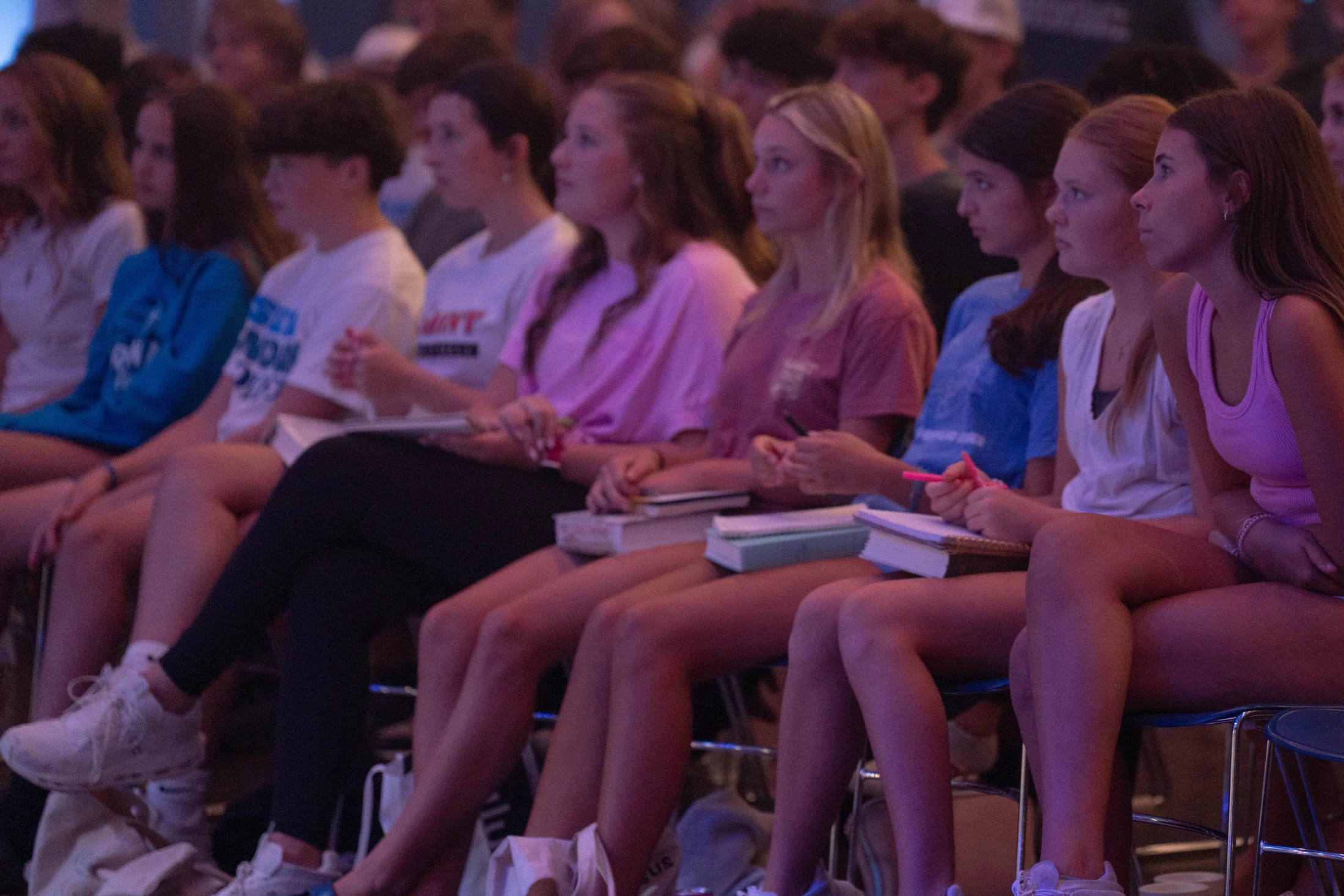 A group of young students, mostly women, sitting in chairs and attentively listening to a presentation, some with notebooks on their laps.