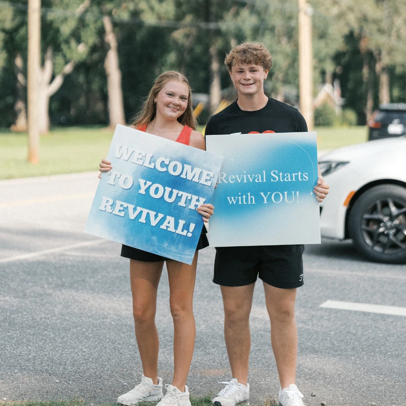 A smiling teenage girl and boy stand outside holding signs that say "Welcome to Youth Revival!" and "Revival Starts with YOU!" next to a road.