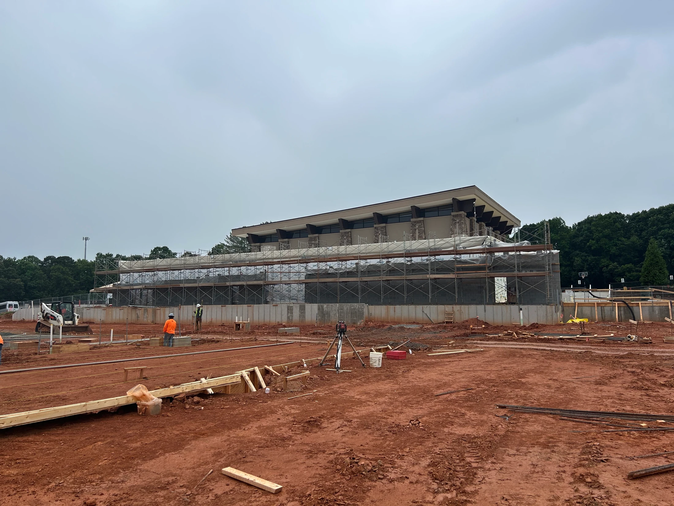 A large construction site with red soil in the foreground and a multi-story modern building surrounded by scaffolding in the background under an overcast sky.