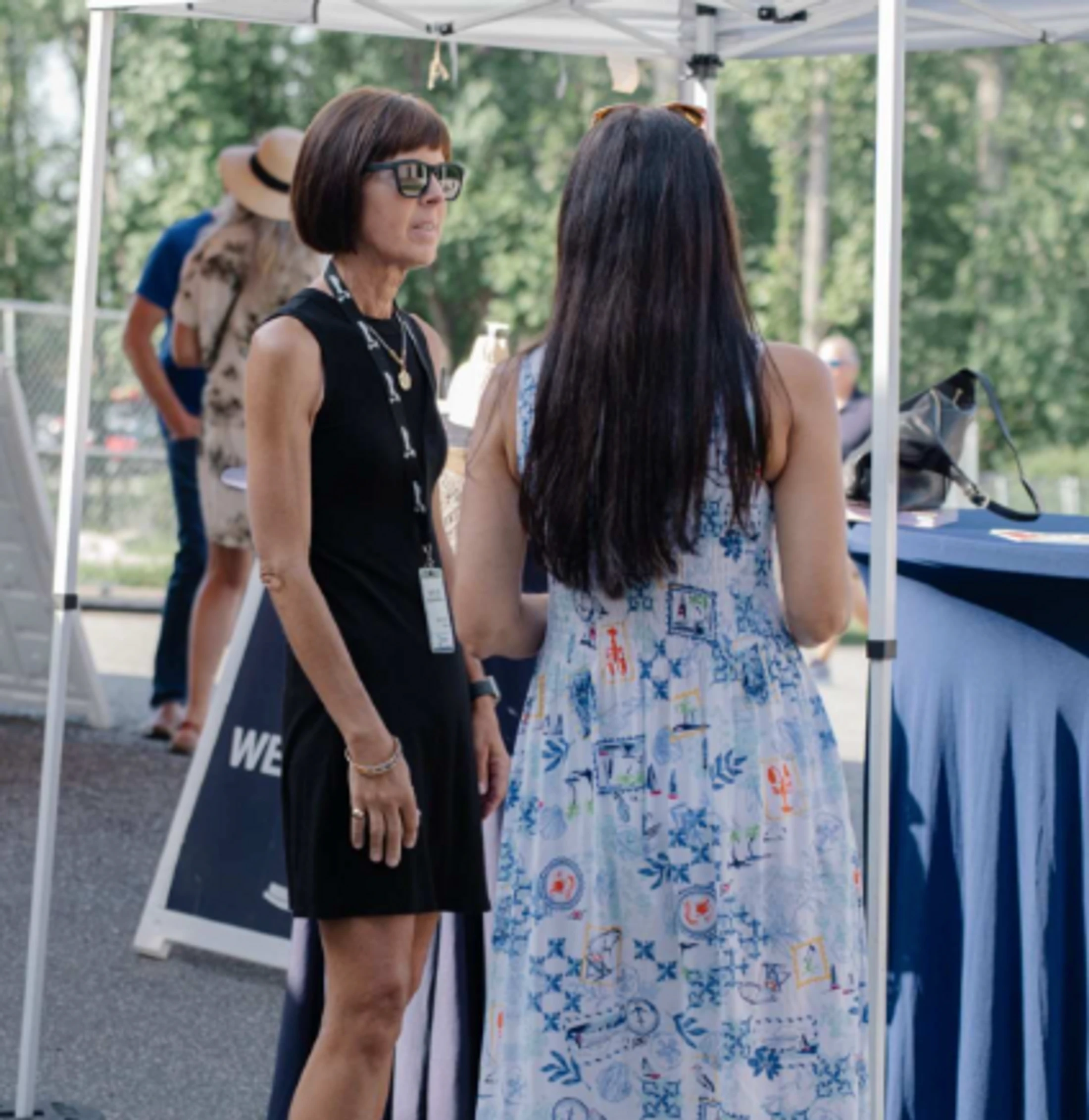A woman in a black dress and sunglasses talks to another woman in a patterned sundress. They are standing under a white canopy tent at an outdoor event.
