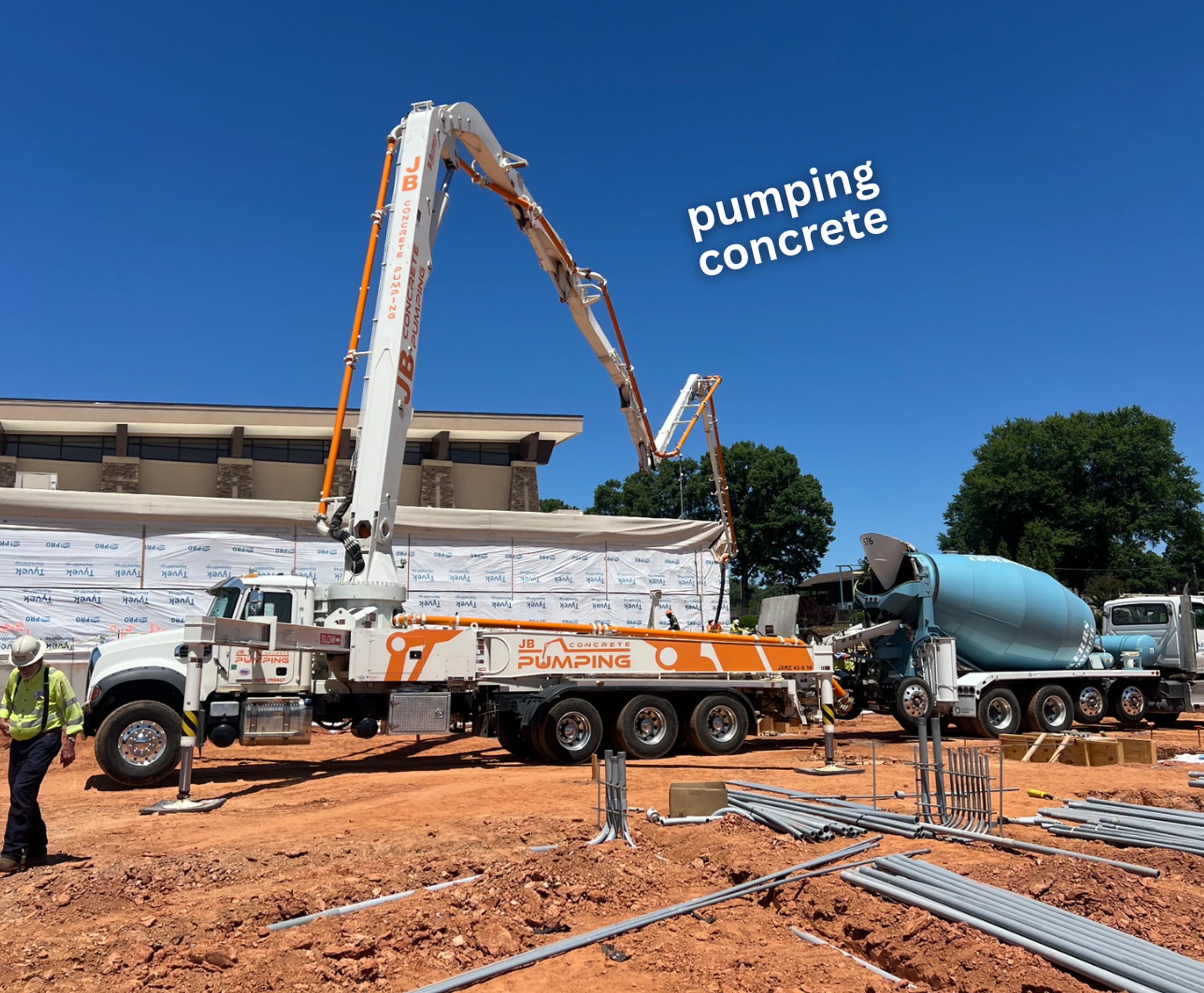 A large concrete pump truck with its boom extended and a concrete mixer truck are on a construction site with red dirt under a clear blue sky.