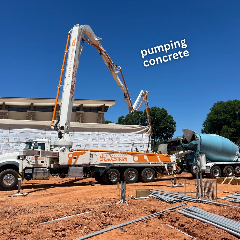 A large concrete pump truck with its boom extended and a concrete mixer truck are on a construction site with red dirt under a clear blue sky.