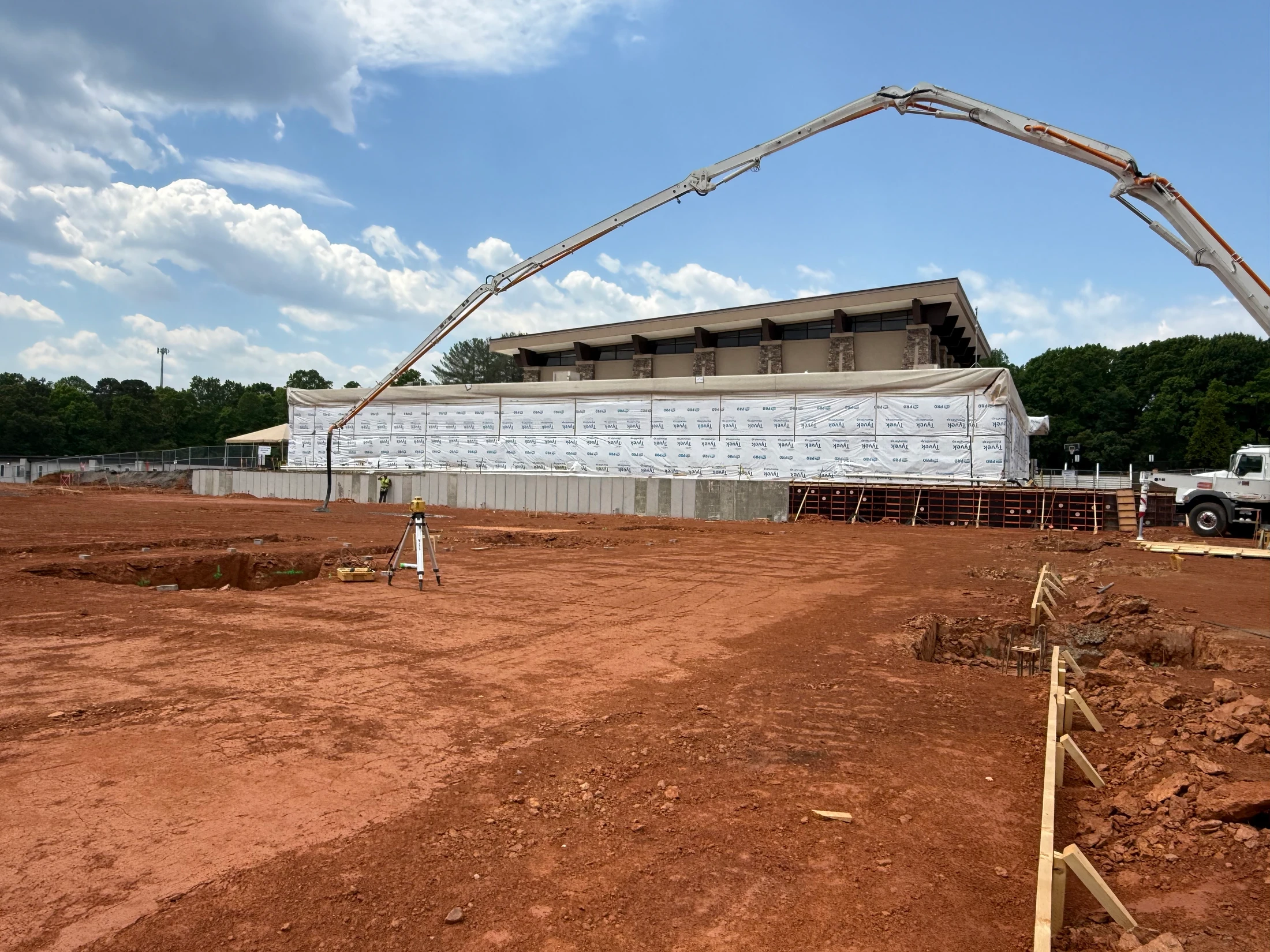 A wide shot of a construction site with red dirt. A large building is being built, and a concrete pump truck has its long boom extended over the site.