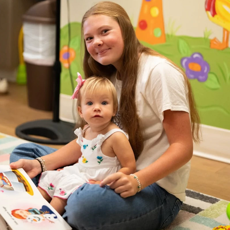 An older girl with long brown hair sits on a colorful rug, holding a baby sister on her lap while they look at a picture book together in a playroom.