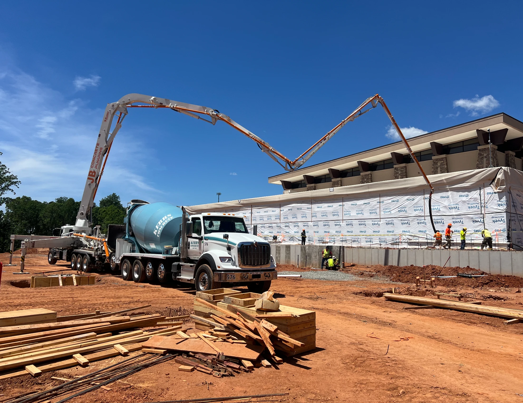 A concrete pump truck with its boom extended over a building foundation, next to a cement mixer truck on a large construction site with workers.