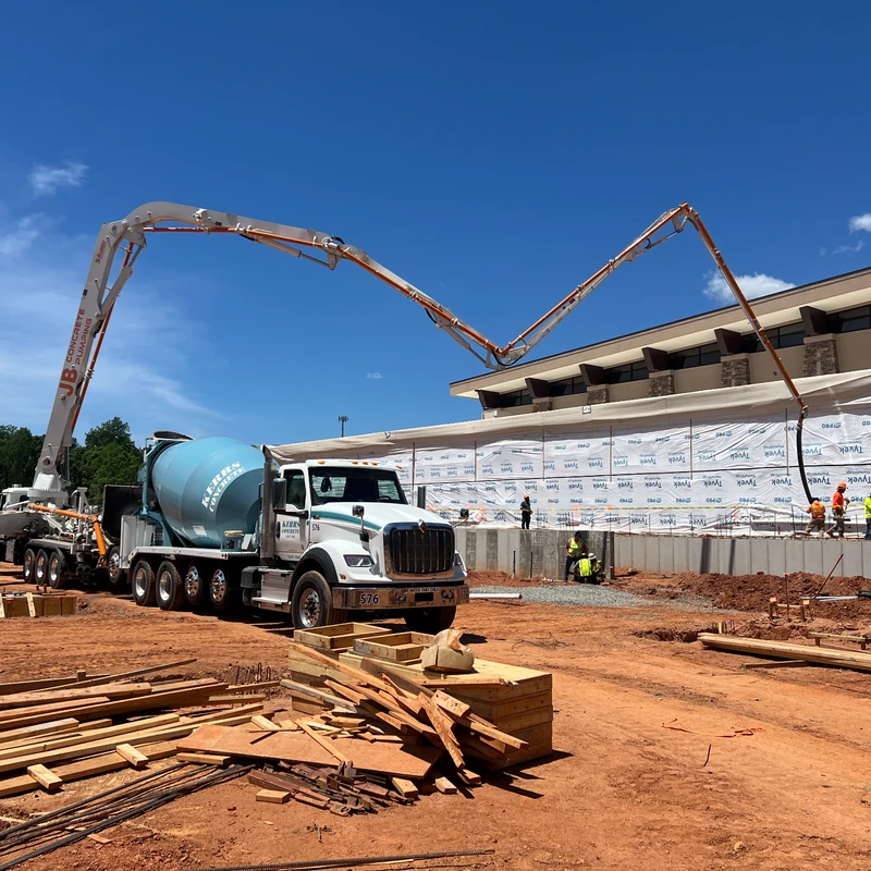 A concrete pump truck with its boom extended over a building foundation, next to a cement mixer truck on a large construction site with workers.