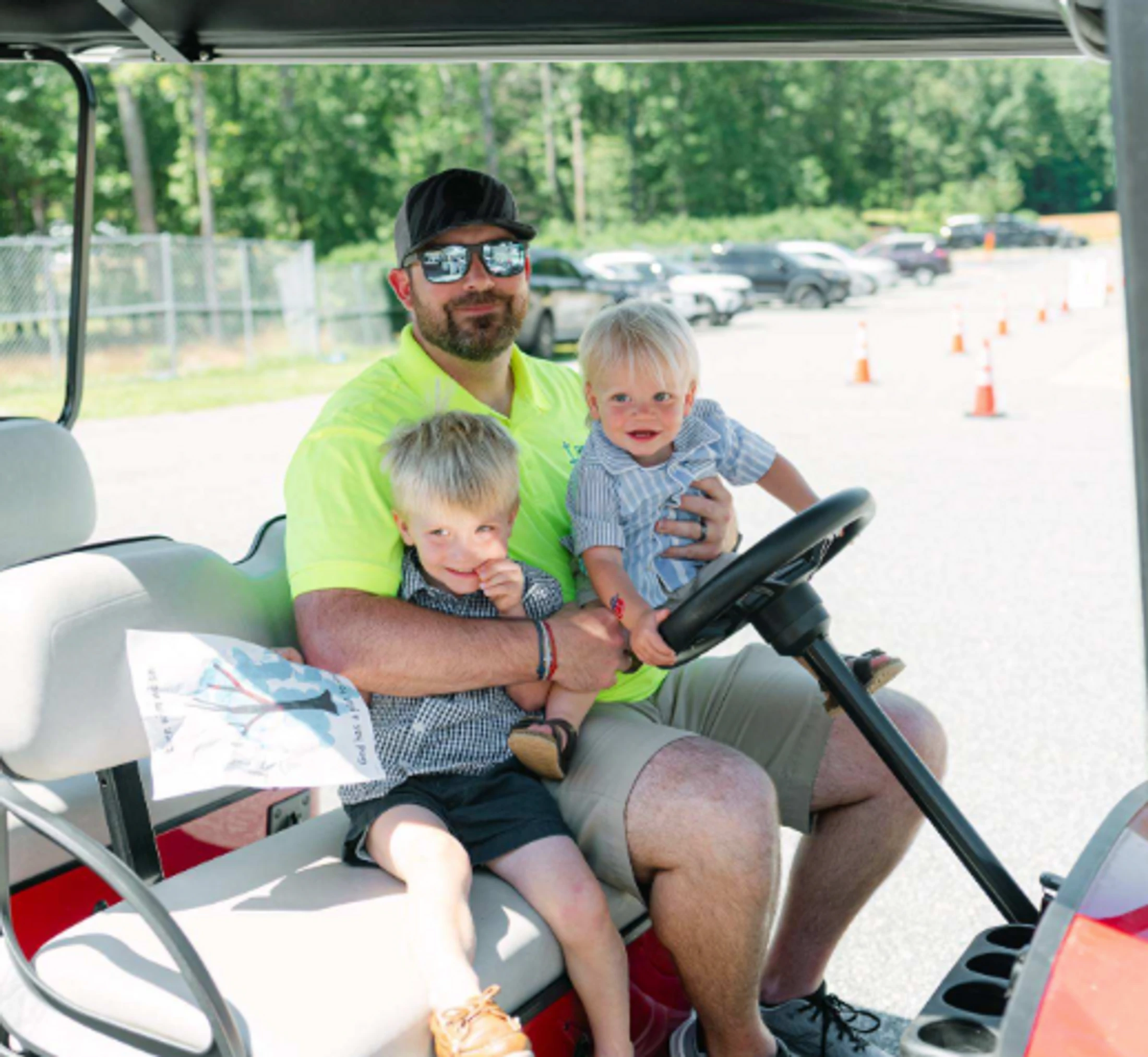A man with a beard and sunglasses sits in a golf cart holding his two young sons on his lap. They are all smiling on a sunny day outdoors.