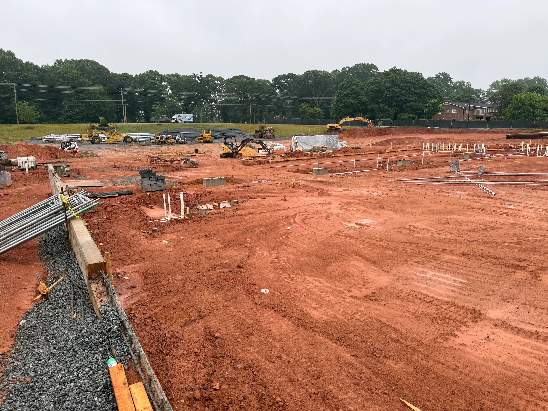 A large construction site with red clay soil. The ground is prepared with plumbing pipes and electrical conduits emerging for a new building's foundation.