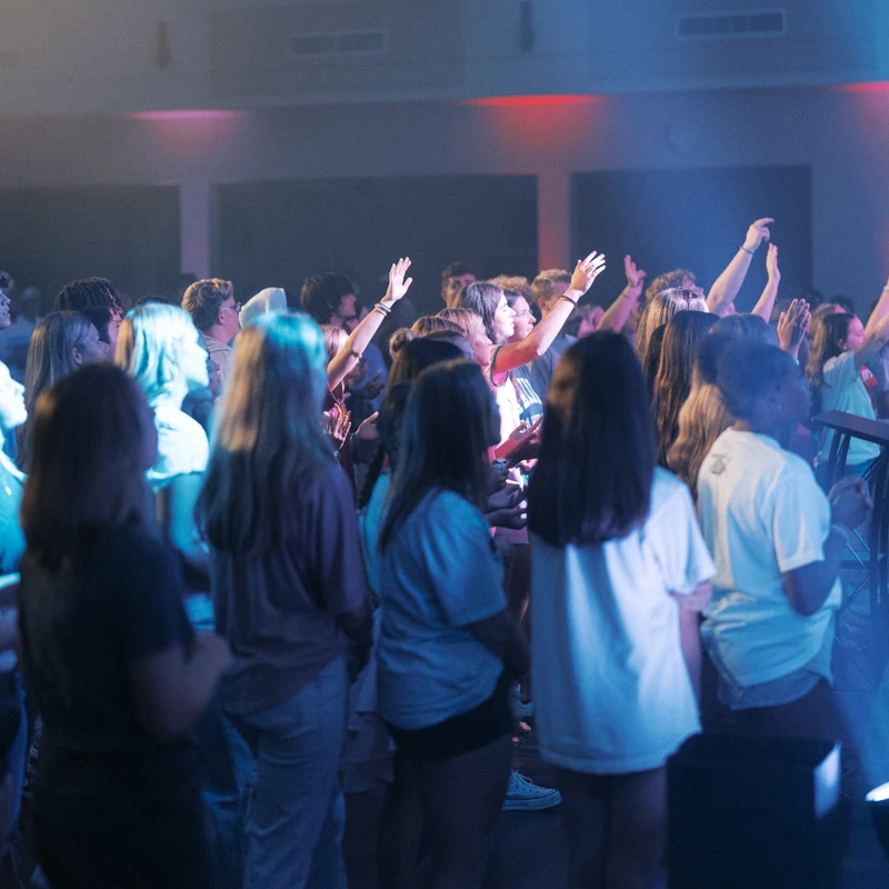 A crowd of young people stand in a dimly lit room with blue and red lights, their hands raised in the air in an act of worship or praise at a concert.
