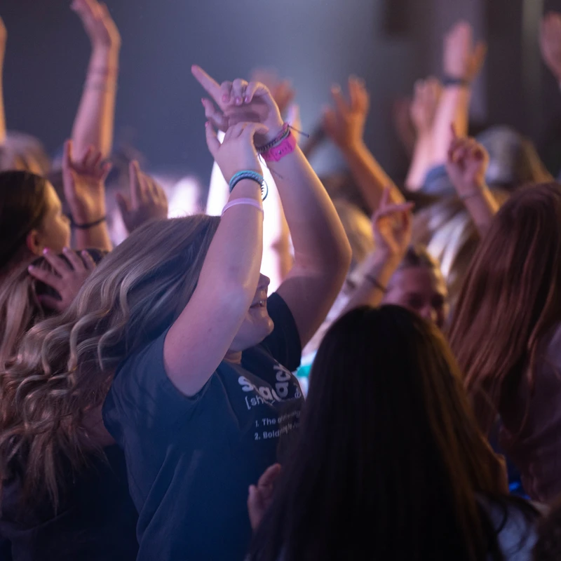A young woman with wavy blonde hair smiles and dances with her arms in the air, surrounded by a large, energetic crowd under purple and white lights.