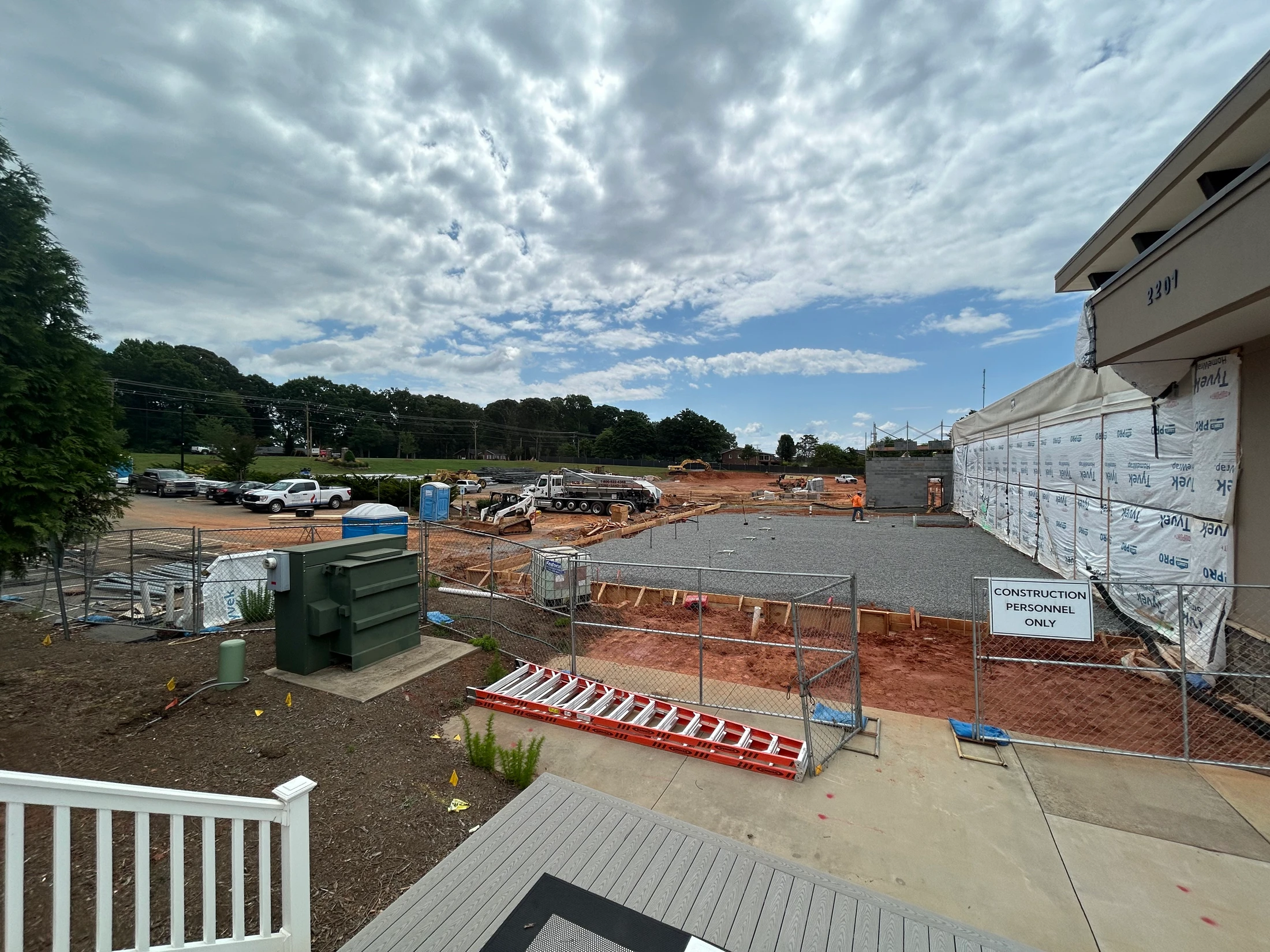 A wide-angle shot of a building construction site with foundations laid, heavy machinery present, and a building wrapped in Tyvek under a cloudy sky.