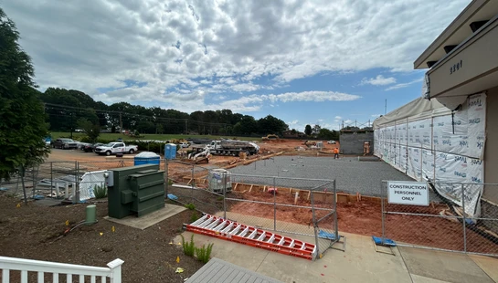 A wide-angle shot of a building construction site with foundations laid, heavy machinery present, and a building wrapped in Tyvek under a cloudy sky.